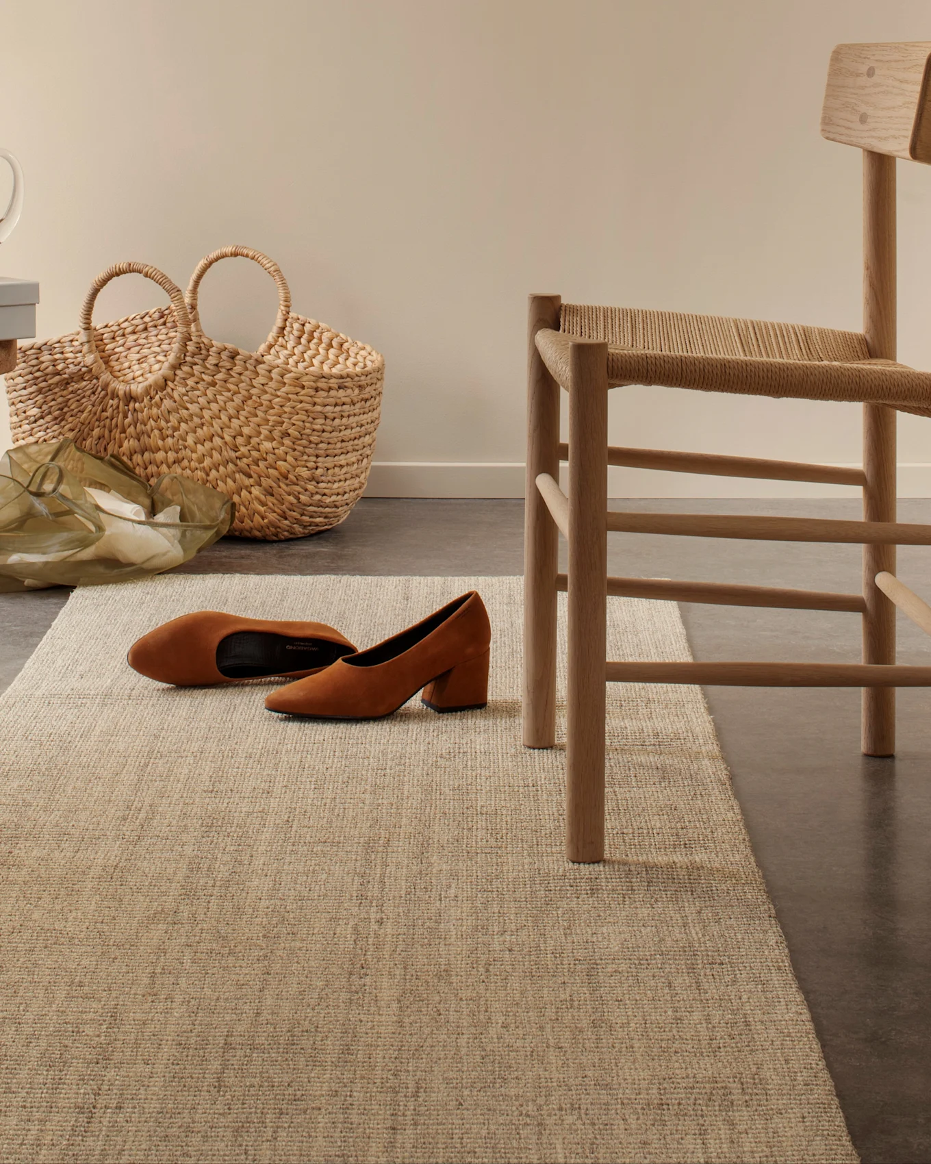 Chaussures en daim marron à talon bloc sur un tapis tissé clair, avec un sac en paille et une chaise en bois en arrière-plan.