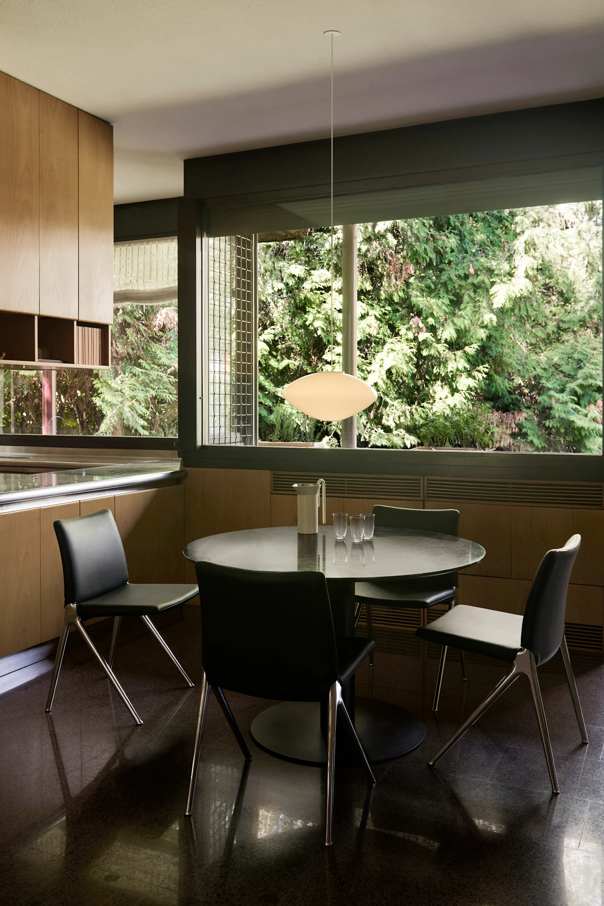 Un coin repas moderne avec une table ronde noire, quatre chaises noires et un luminaire suspendu ovale blanc. Des armoires en bois clair bordent un mur, avec de grandes fenêtres donnant sur des arbres verts.