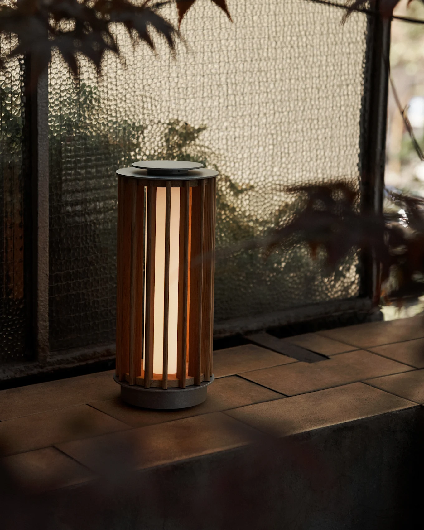 Une lampe portable cylindrique avec un corps en bois à lattes et une lueur chaude, sur une surface carrelée près d'une fenêtre en verre texturé.