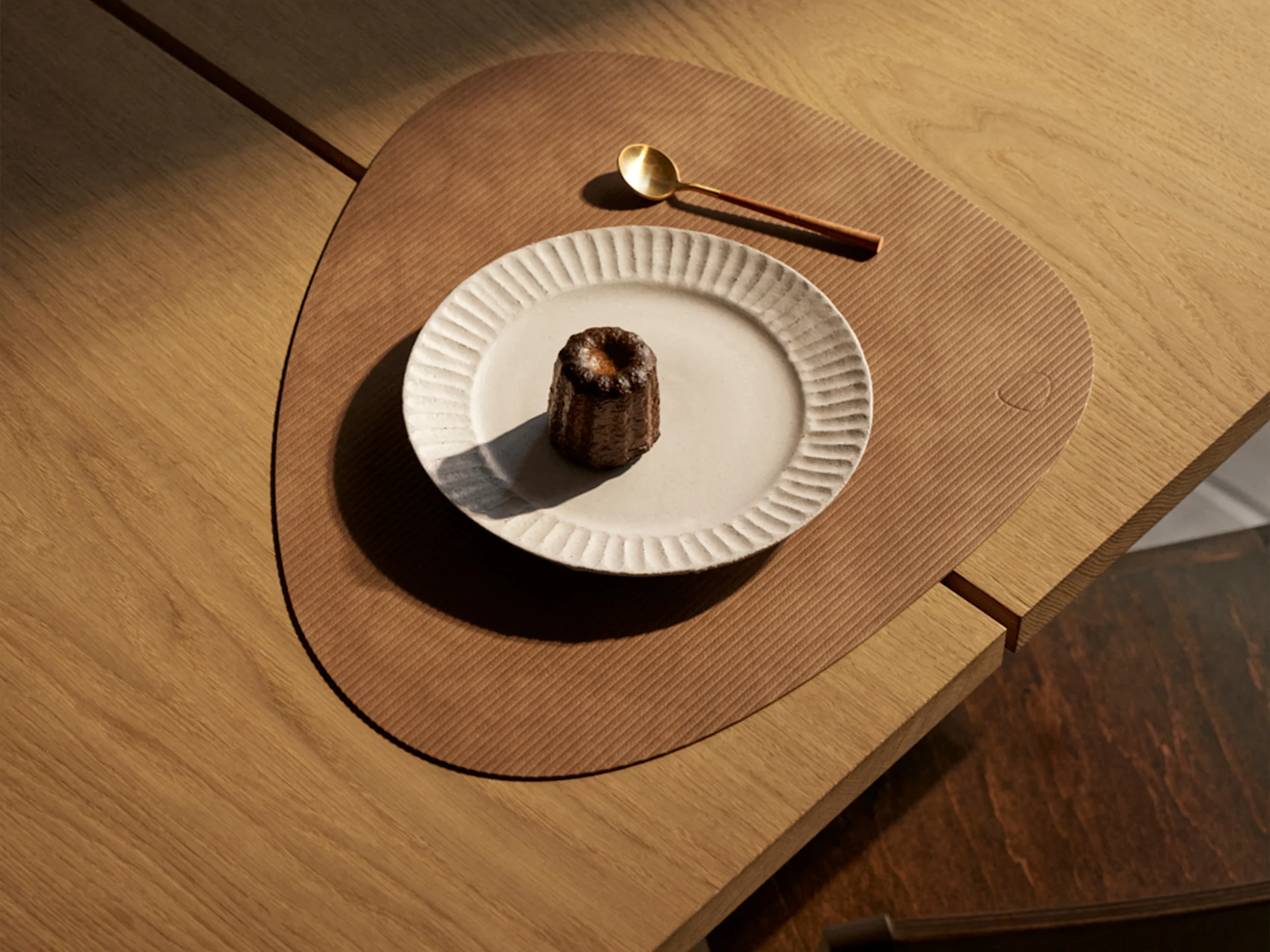 Un canelé sur une assiette nervurée, une cuillère dorée et un set de table triangulaire sur une table en bois, éclairé par le soleil.
