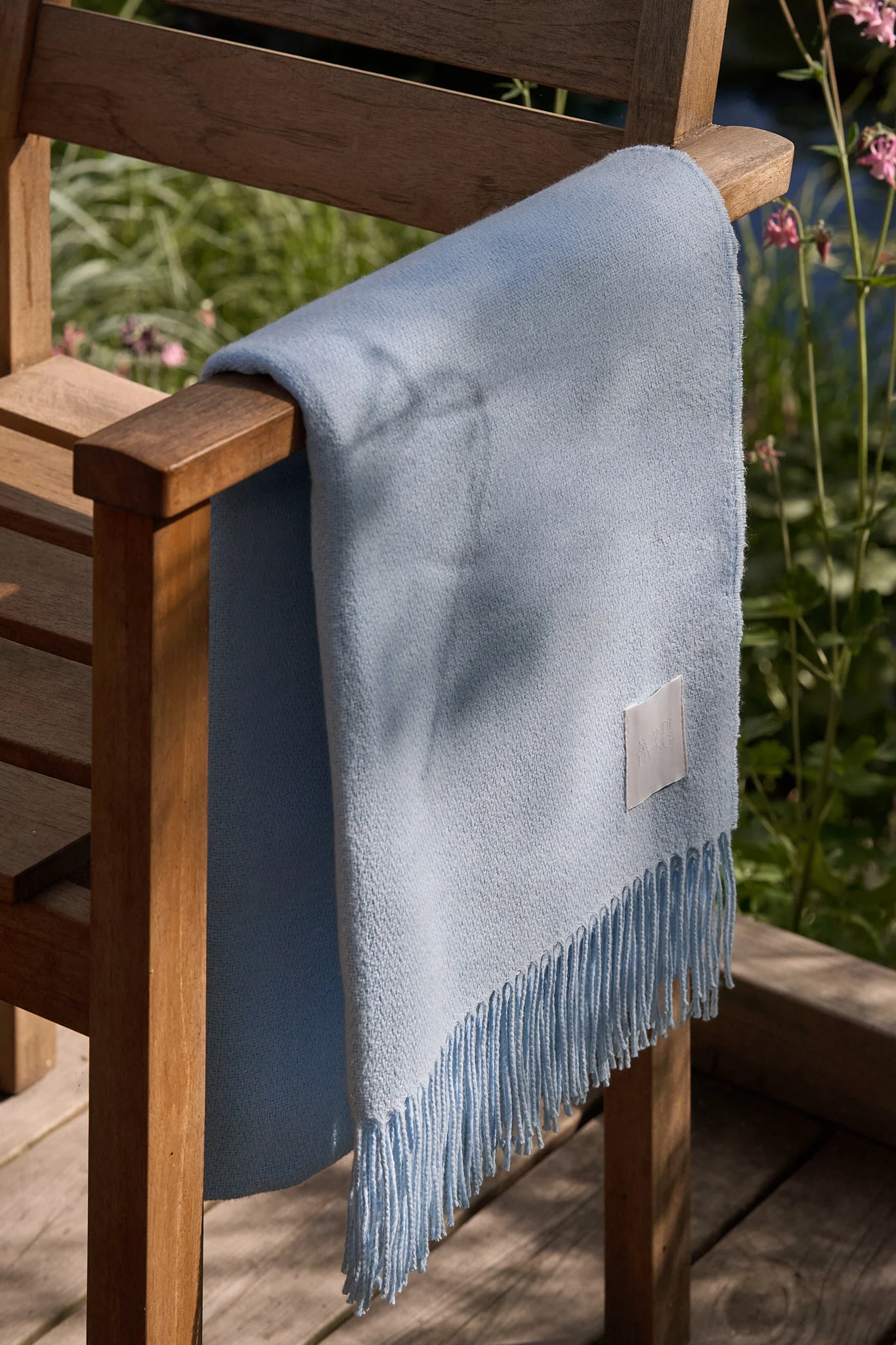 Couverture bleu clair à franges drapée sur une chaise de jardin en bois dans un jardin ensoleillé.