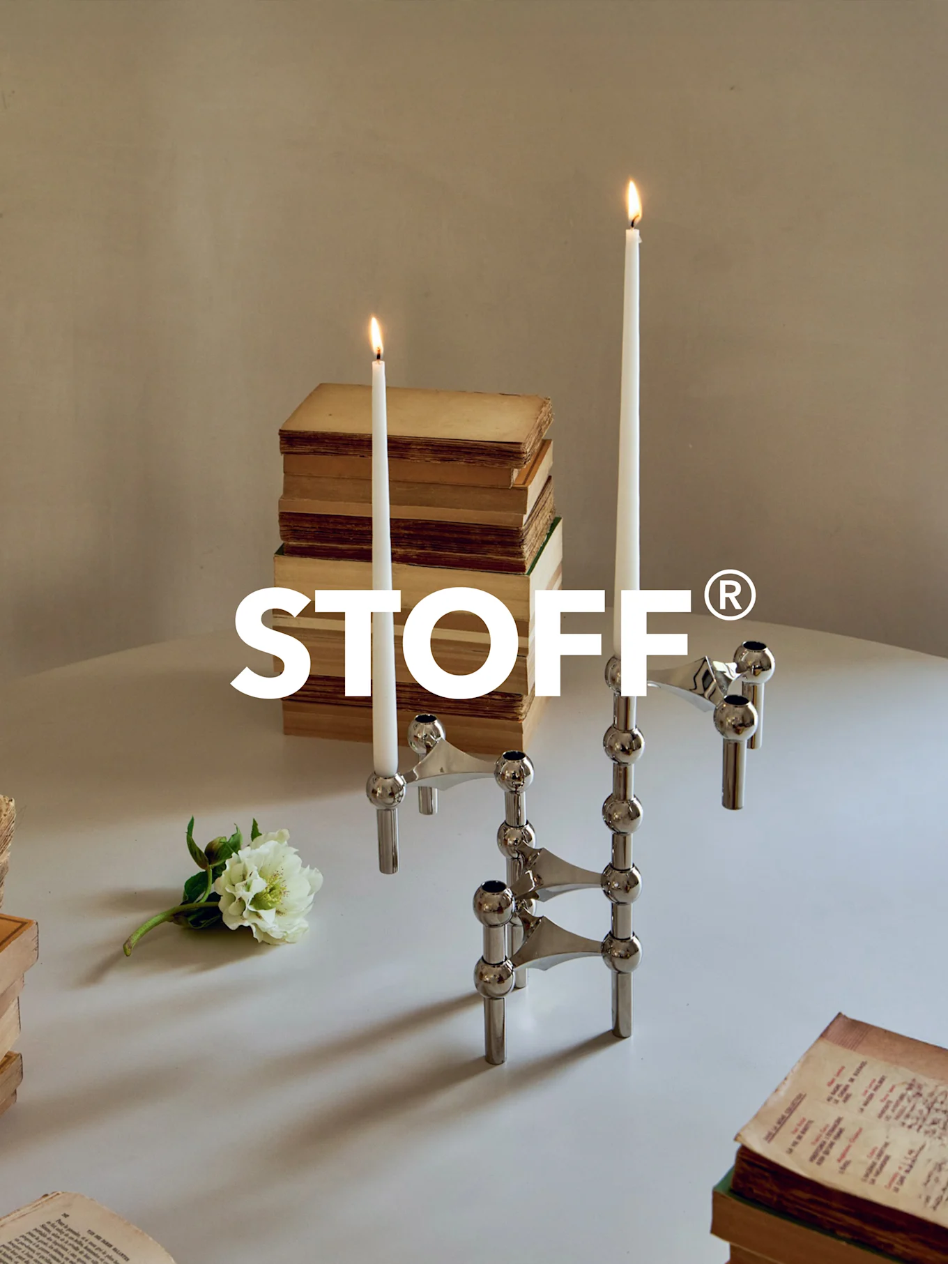 Candélabre modulaire en chrome poli avec deux bougies allumées, entouré de vieux livres et d'une fleur blanche sur une table. Logo STOFF®.