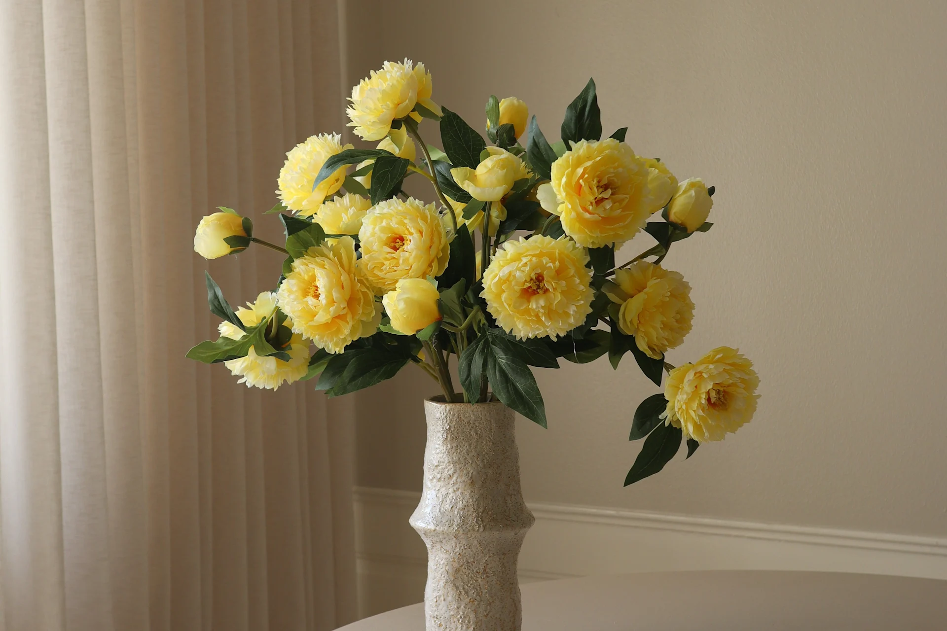 Un vase clair et texturé contient un bouquet éclatant de fleurs jaunes de type pivoine et de feuilles vertes sur un fond clair.