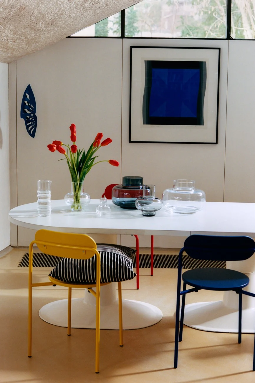 Salle à manger moderne avec table ovale blanche, tulipes rouges, vases en verre, chaises jaunes et bleues, et art abstrait bleu.