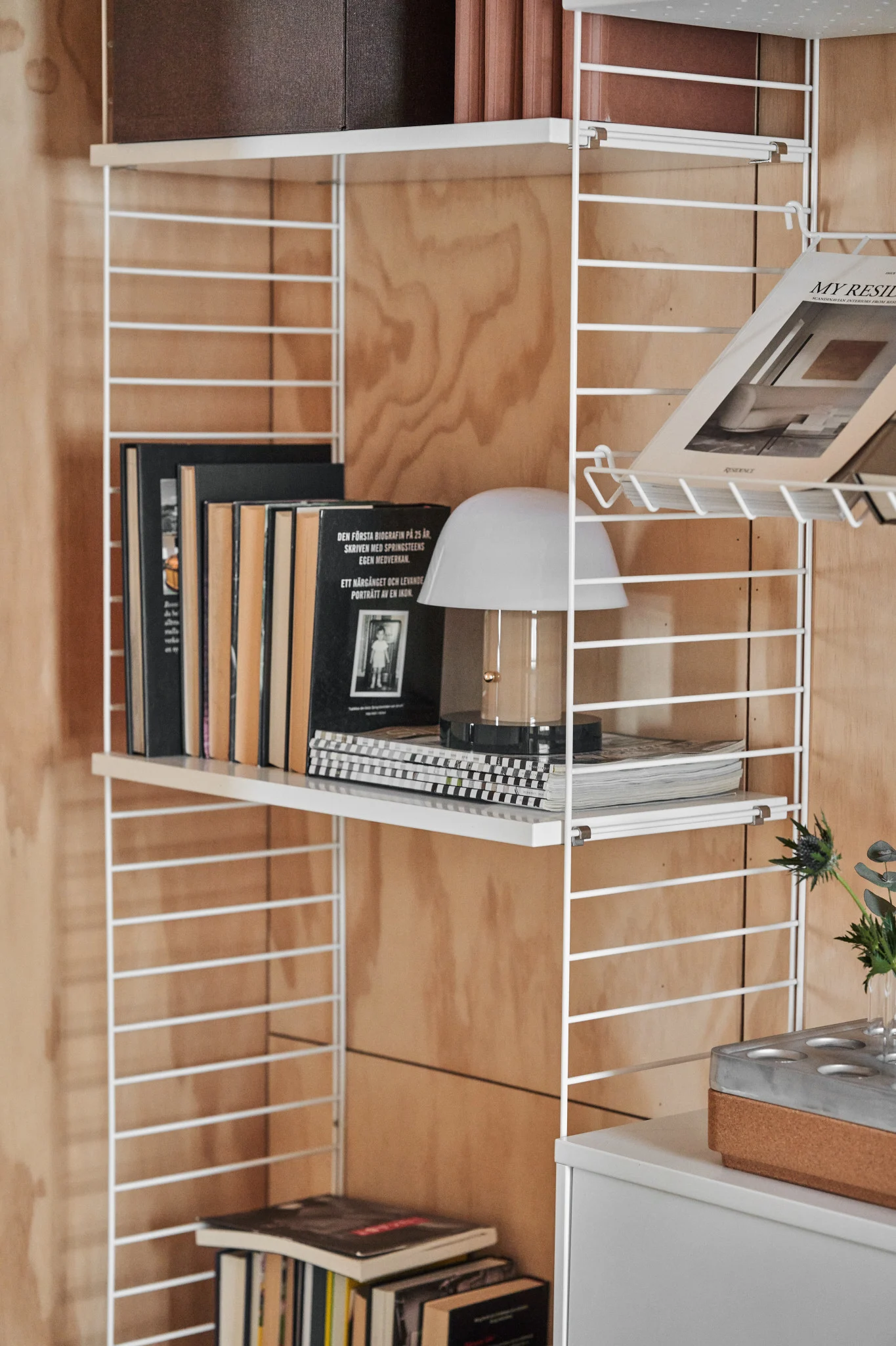 Étagère murale en fil métallique blanc sur un mur en bois, présentant des livres, une lampe de table blanche et un magazine. Un meuble gris est en dessous.