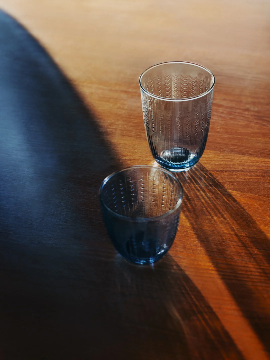 Deux verres texturés teintés de bleu sur une table en bois, avec de longues ombres.