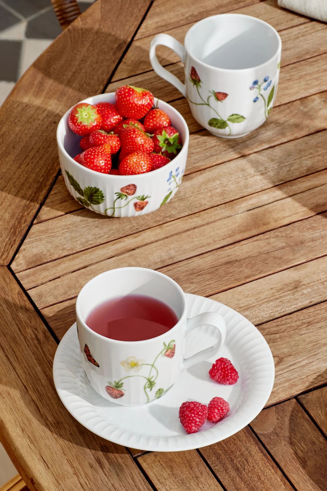 Une tasse blanche avec du thé rose et une soucoupe avec des framboises, un bol de fraises et une tasse vide sur une table en bois sous le soleil.