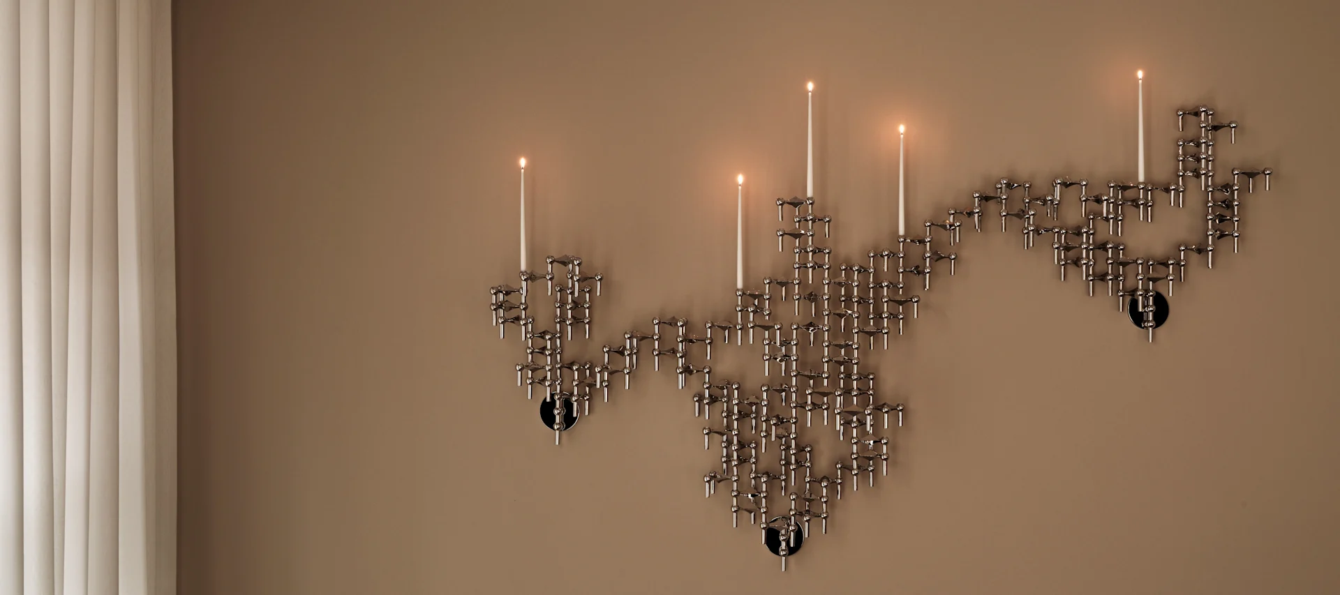 Un candélabre modulaire argenté fixé au mur, sur lequel brûlent plusieurs bougies, devant un mur beige, à côté de stores verticaux.