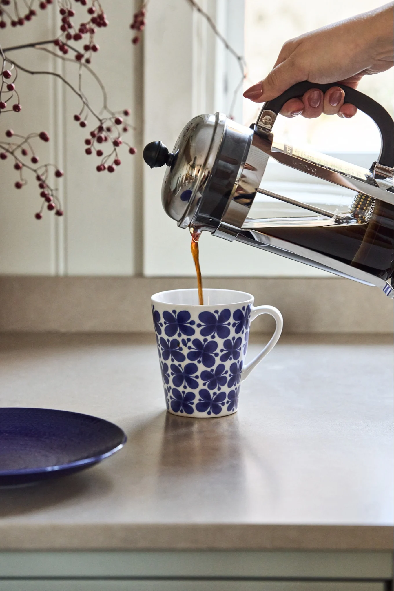 Une main verse du café d'une cafetière à piston dans une tasse blanche à motifs floraux bleus sur un comptoir de cuisine.