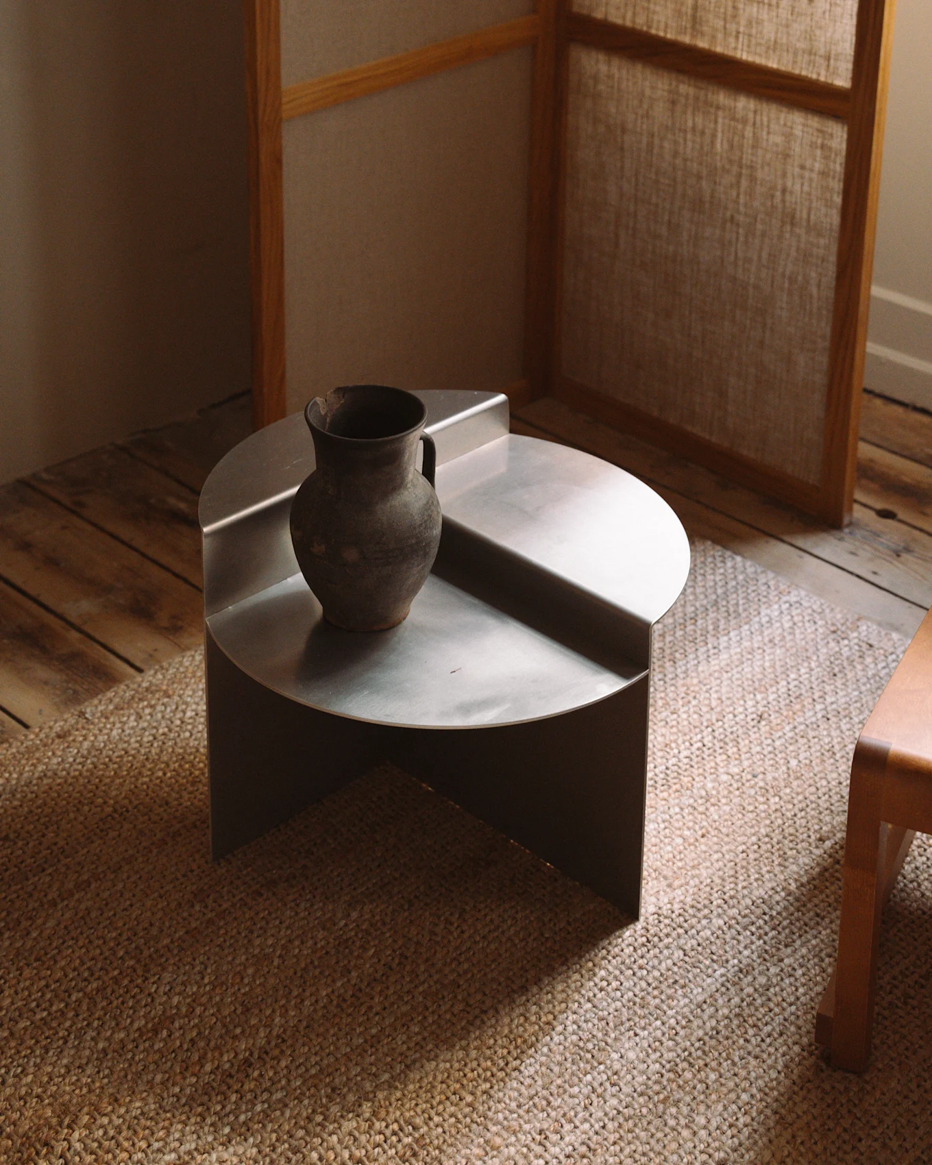 Une table basse minimaliste, ronde et basse en métal argenté avec un vase rustique en céramique foncée sur un tapis tissé.