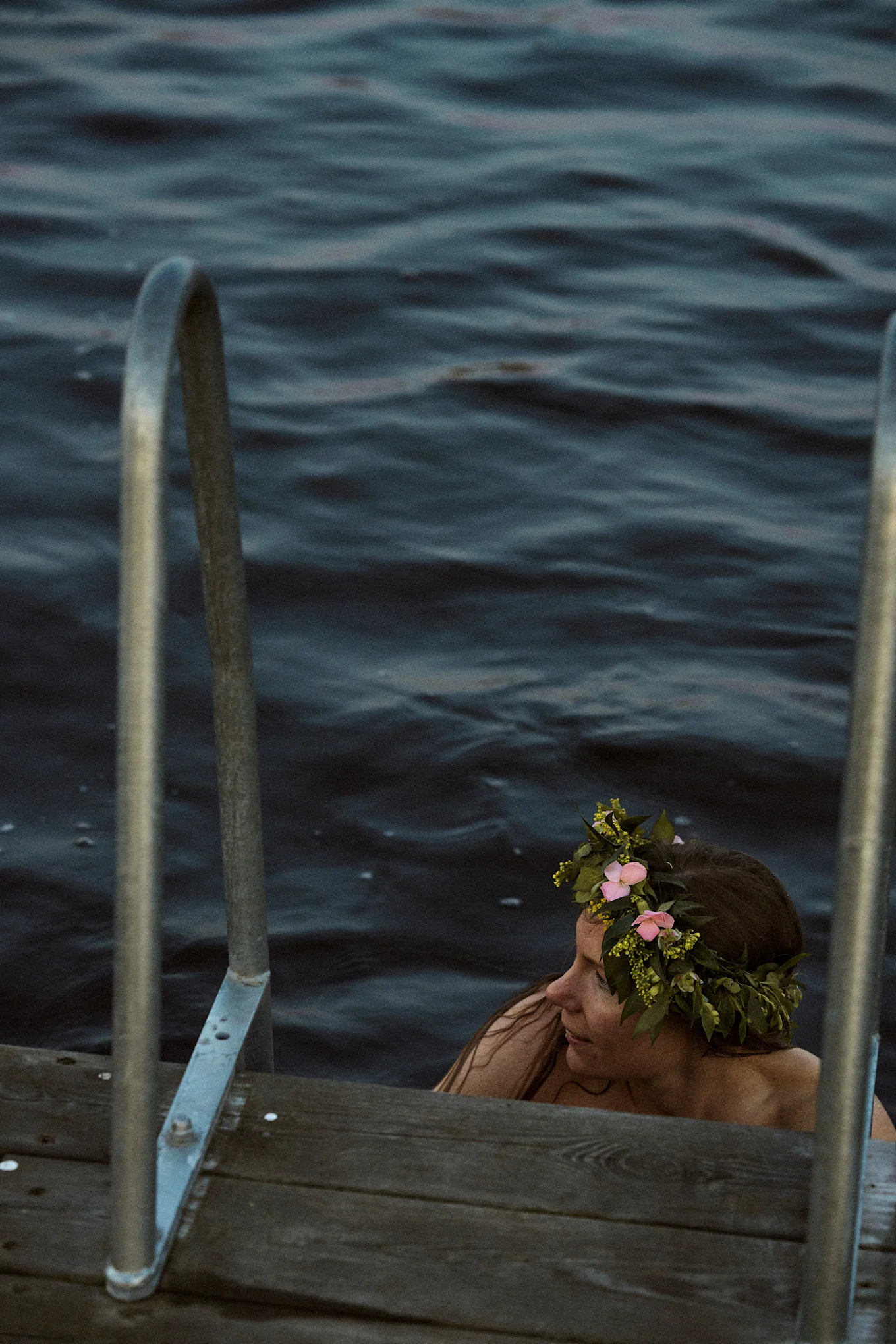 Femme dans l'eau sombre avec une couronne de fleurs, près d'un quai en bois avec des mains courantes métalliques.