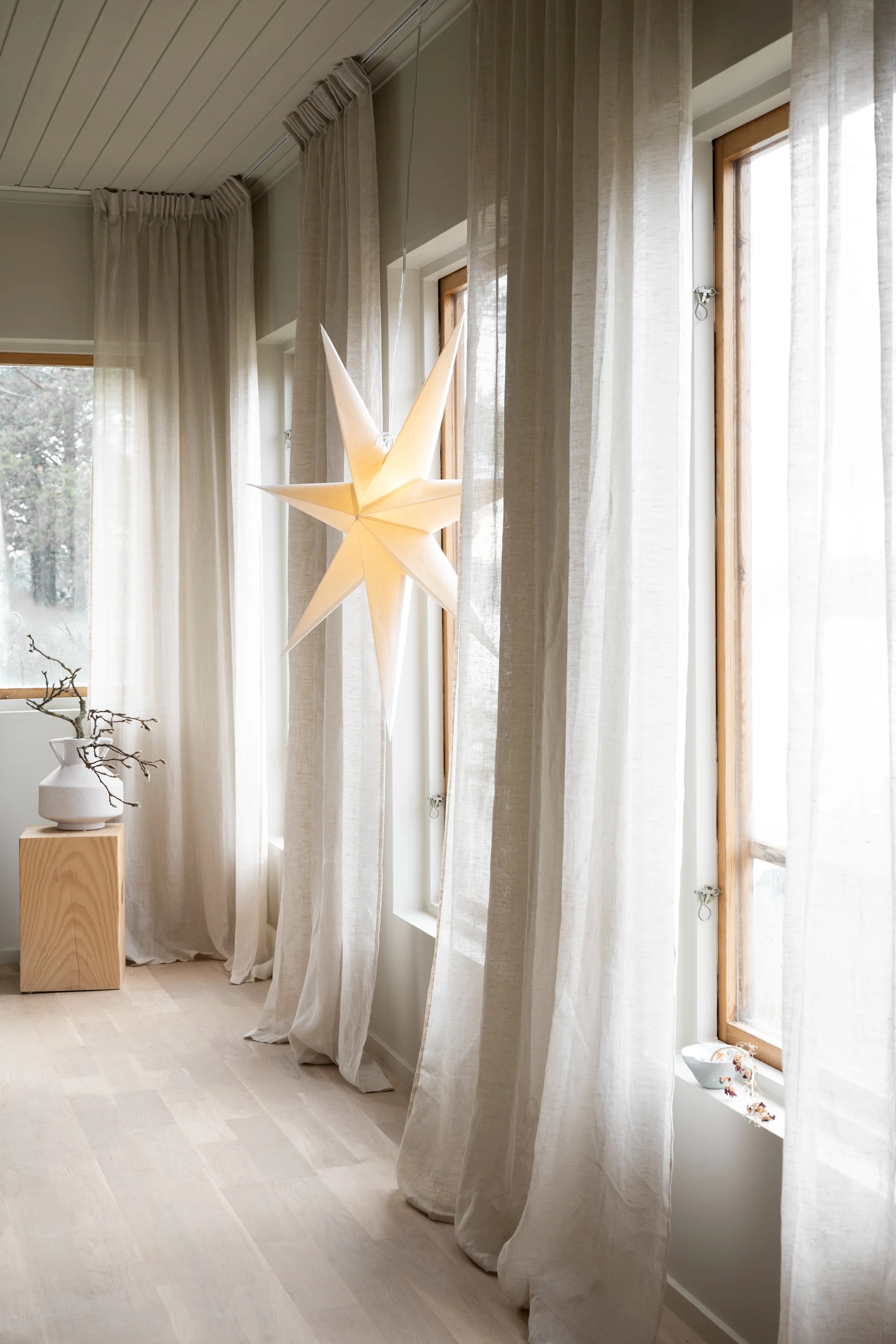 Une étoile en papier illuminée est suspendue dans une fenêtre avec des rideaux transparents, à côté d'un vase blanc avec des branches sur un bloc de bois.