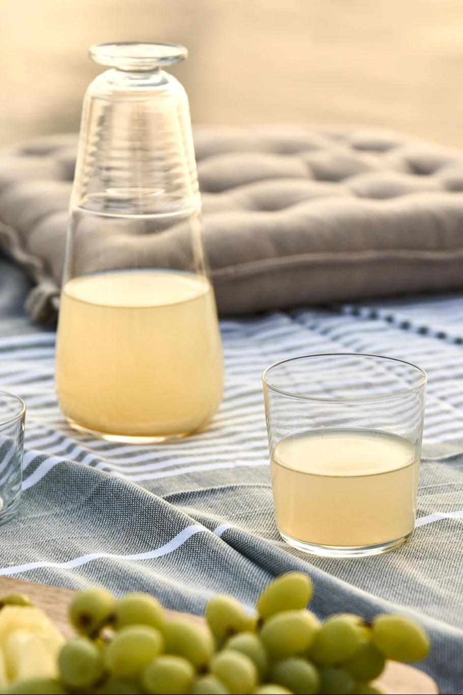 Découvrez des recettes estivales simples : vous voyez ici une bouteille de jus de citron avec un verre et une grappe de raisin disposés sur une table de pique-nique.