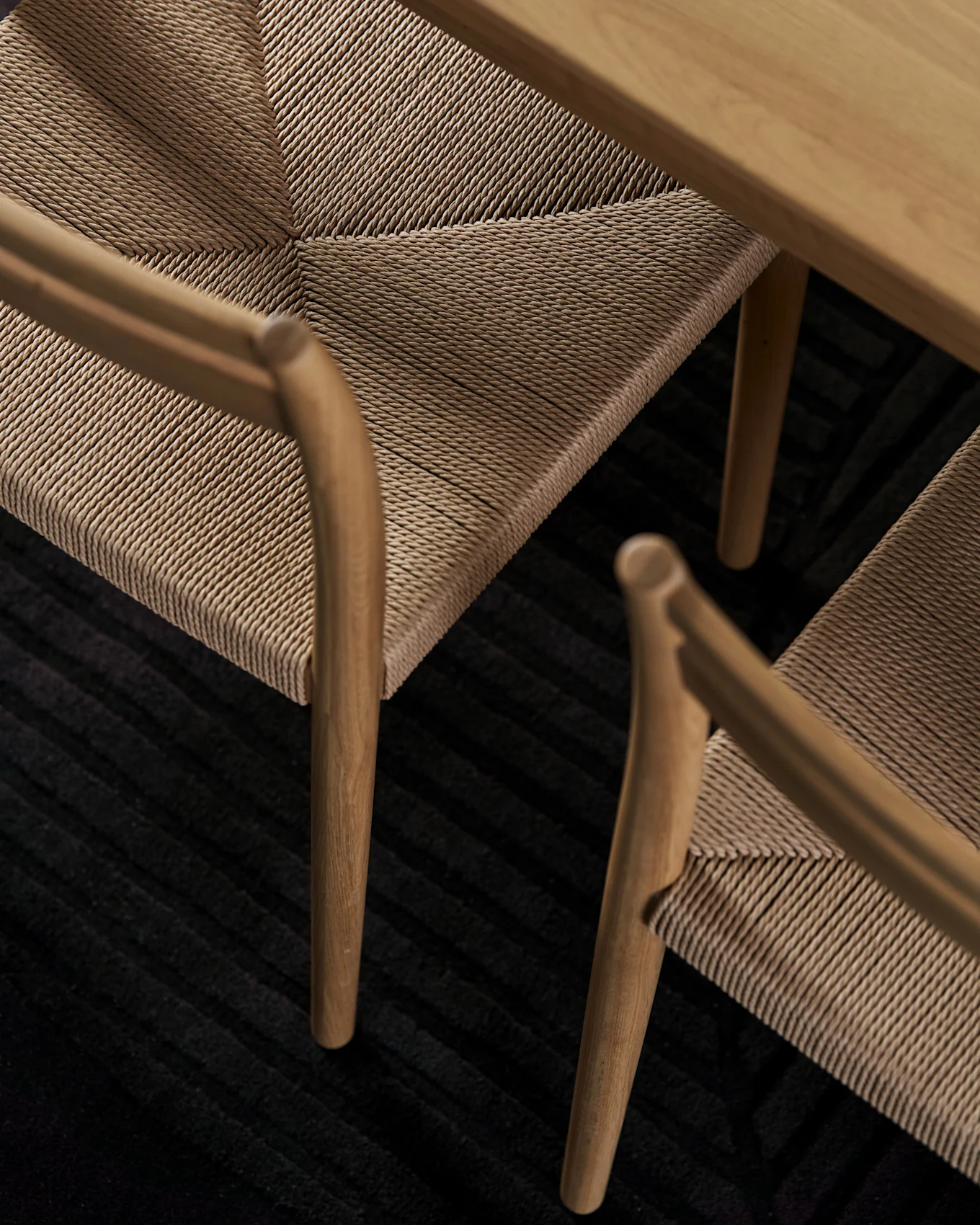 Gros plan de chaises en bois avec des sièges en corde tressée sous une table en bois sur un tapis sombre nervuré.