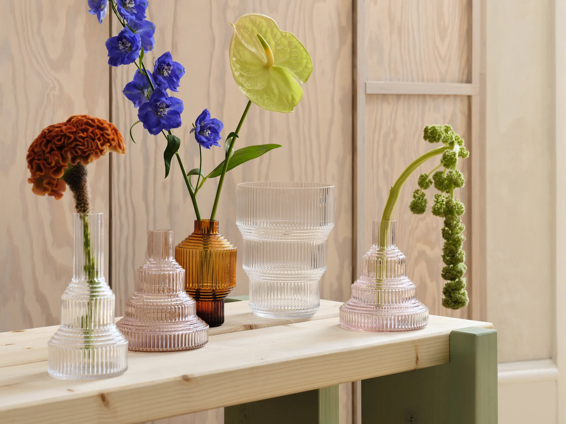 Assortiment de vases en verre côtelé transparent, rose et ambre avec diverses fleurs sur une table en bois clair.