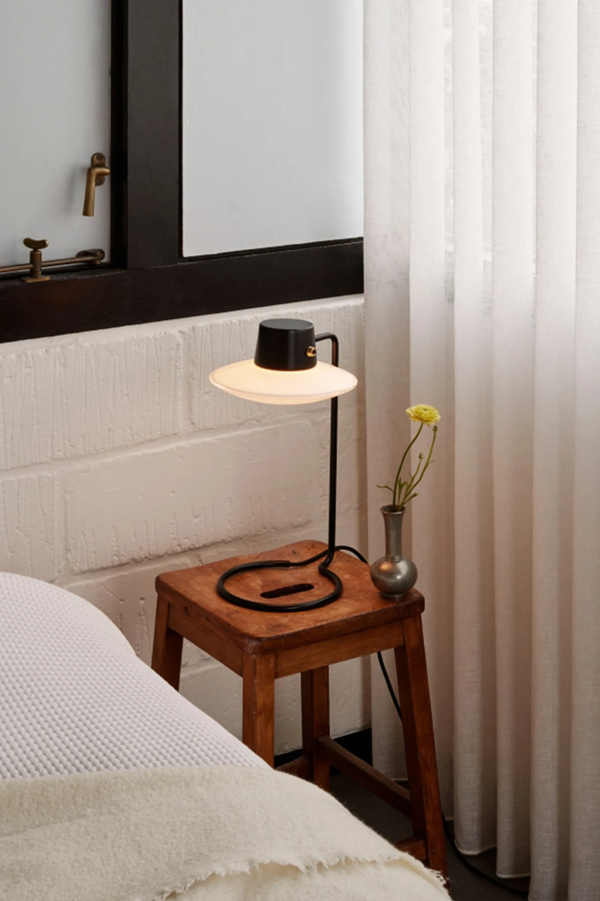 Lampe de table noire à abat-jour large sur tabouret en bois rustique, vase avec fleur jaune, près d'un lit et rideaux.