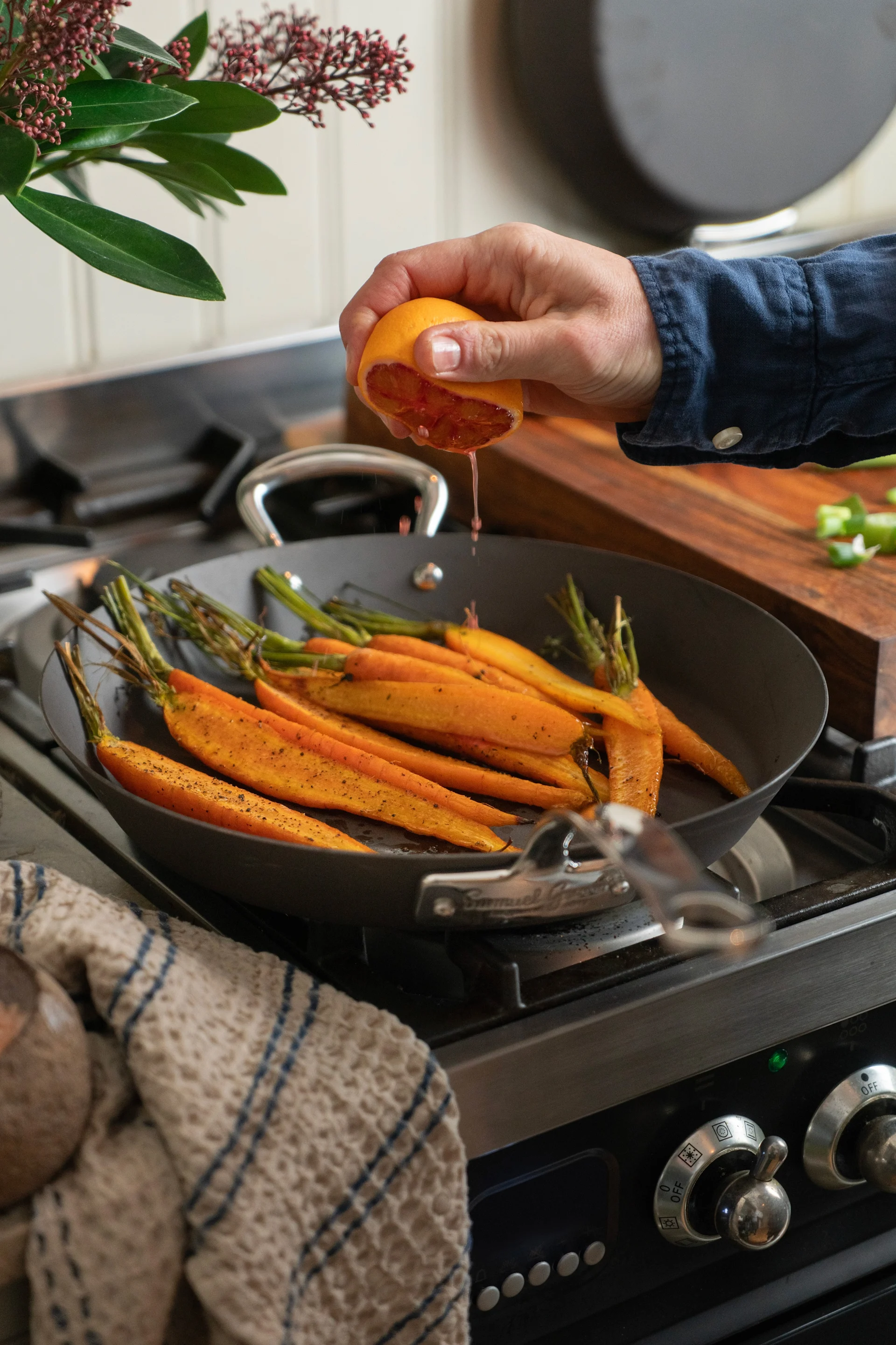 Une poêle en acier au carbone avec des carottes est posée sur la cuisinière, tandis qu'une main presse le jus d'un agrume dessus.