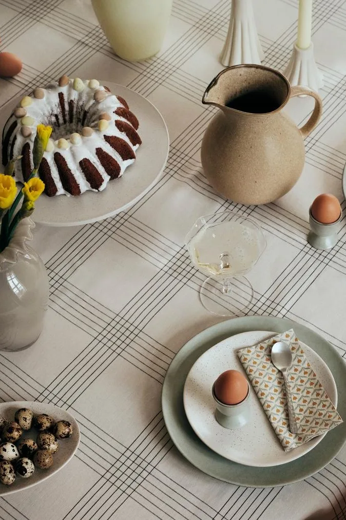 Table de Pâques avec un gâteau, des jonquilles, des œufs et des boissons sur une nappe quadrillée.