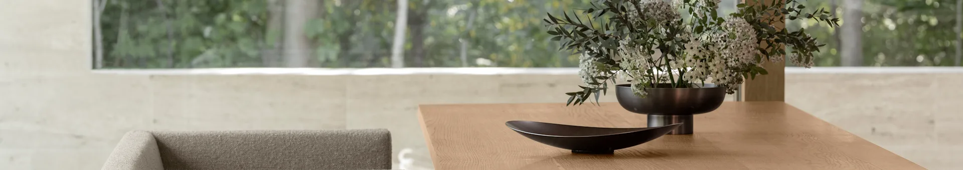 Table à manger moderne en bois avec un bol décoratif foncé et un vase de fleurs blanches et vertes, avec une vue extérieure verte floue à travers une grande fenêtre.