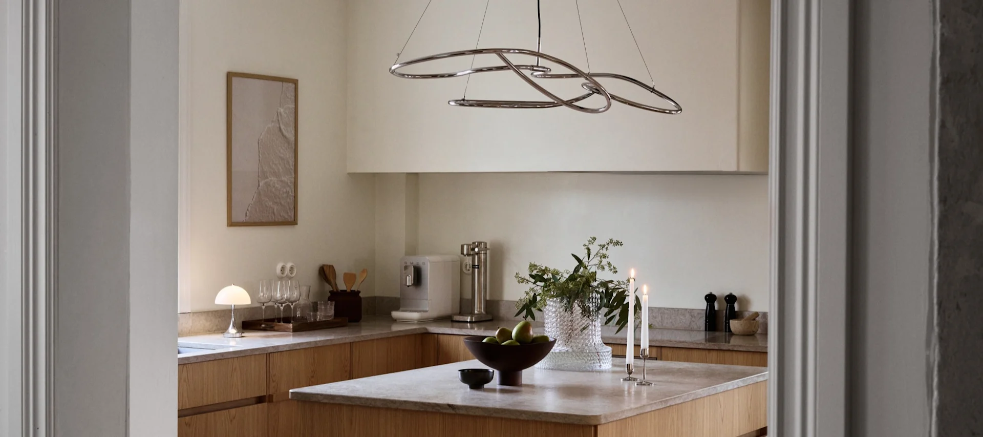 Cuisine moderne avec armoires en bois clair, îlot avec pommes vertes, bougies allumées et lustre sculptural chromé.