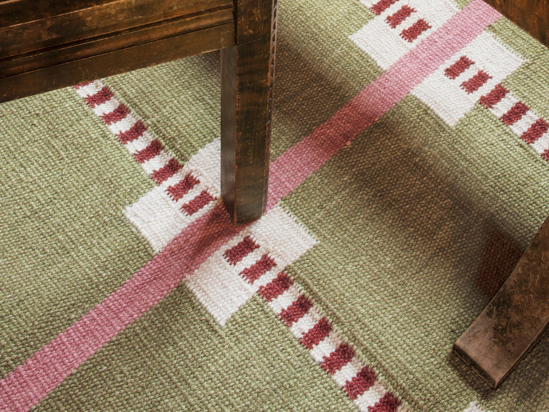 Un tapis à motifs géométriques verts, blancs et rouges avec un pied de meuble en bois foncé projetant une ombre.