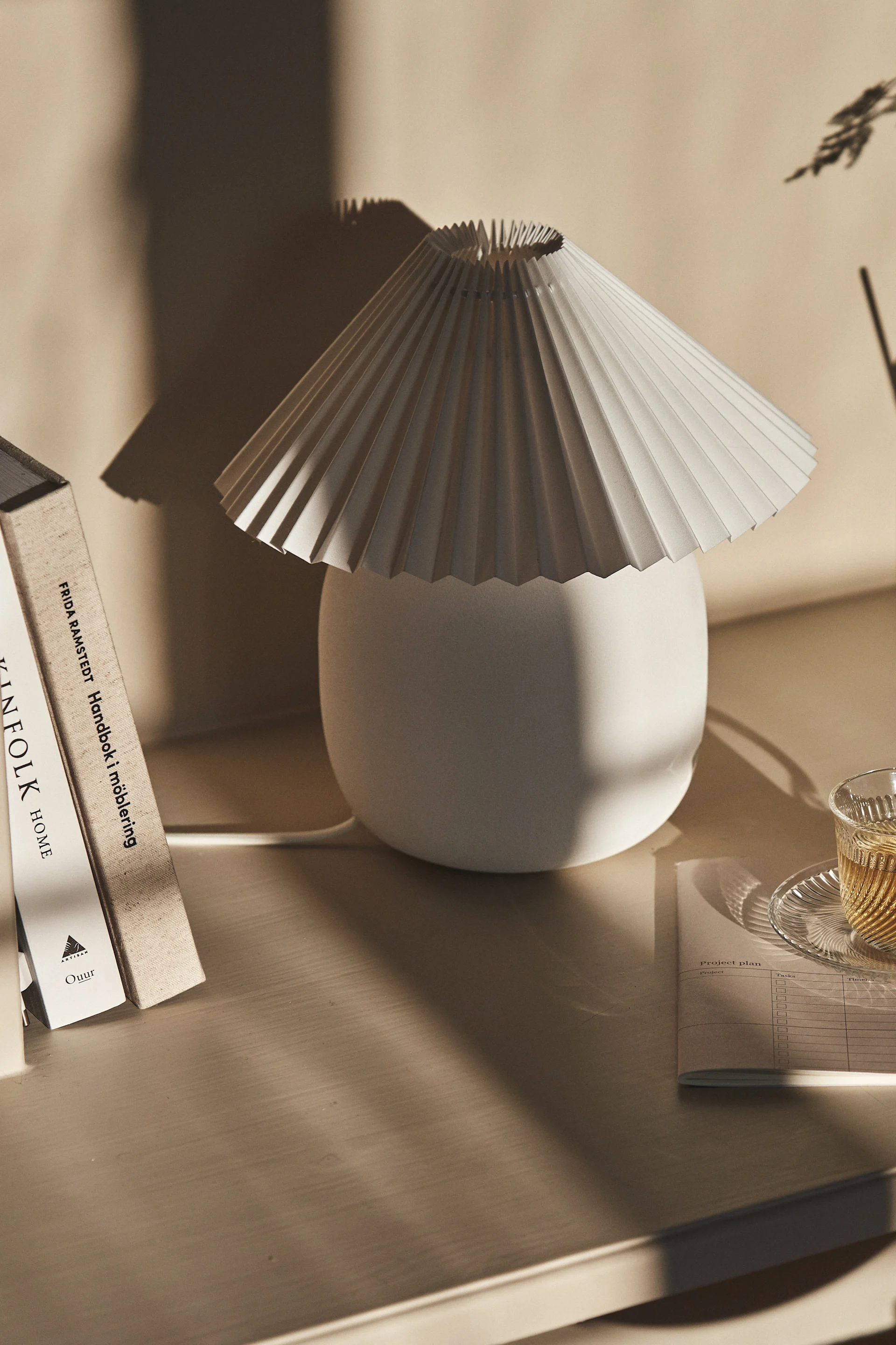 Une lampe de table blanche plissée à côté de livres et d'une tasse de thé sur un bureau en bois clair, avec des ombres douces.