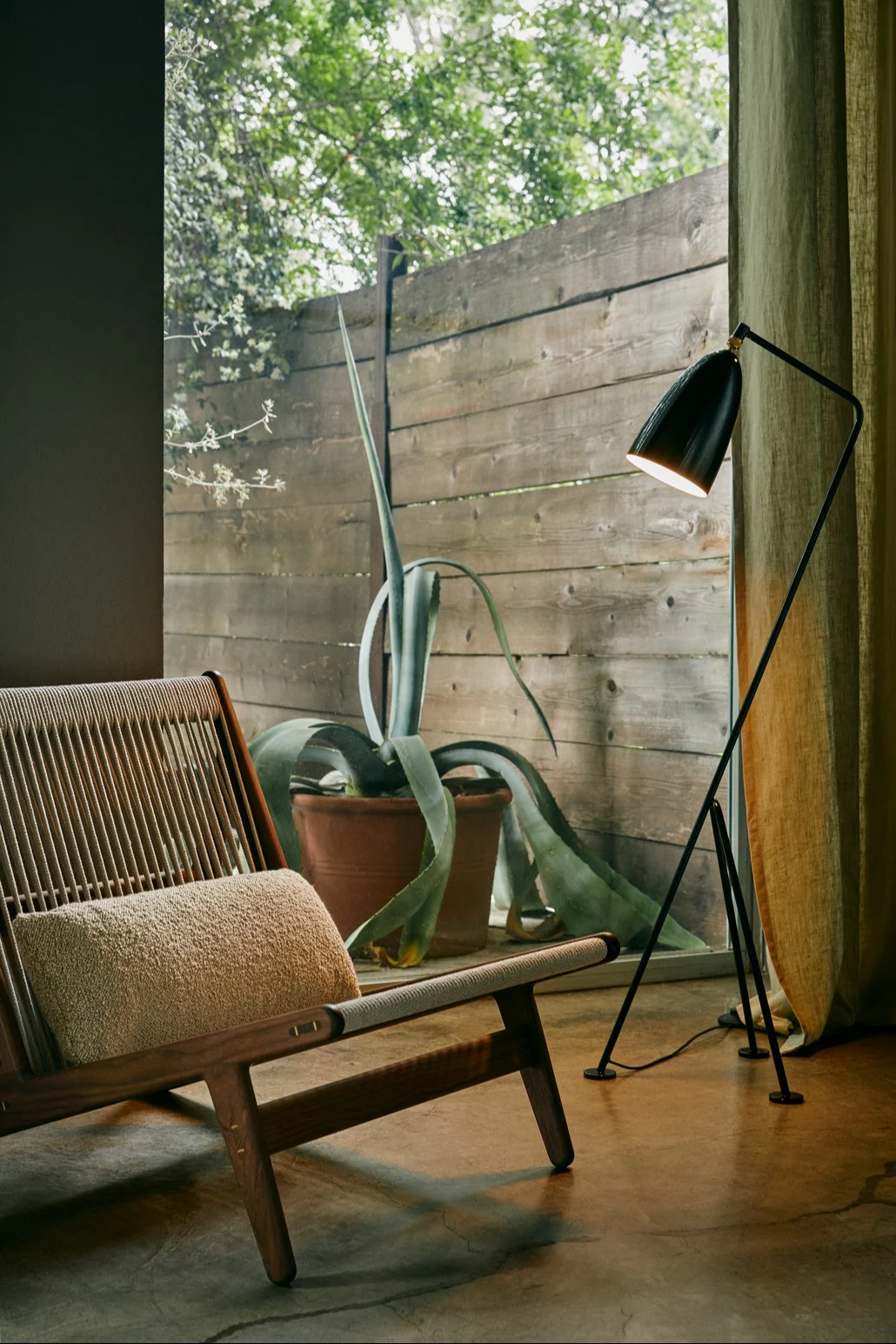 Le lampadaire noir scandinave Gräshoppa de Gubi est placé à côté d'un fauteuil lounge devant une grande fenêtre.