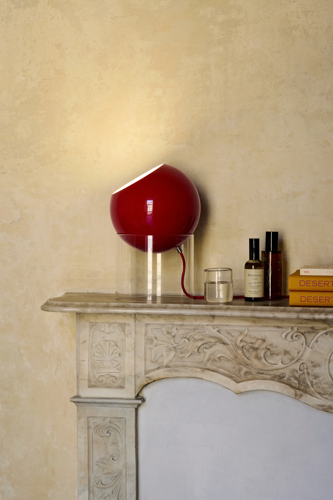 Lampe sphérique rouge avec base translucide sur cheminée en marbre décorée, à côté d'un verre d'eau, deux bouteilles et deux livres jaunes.