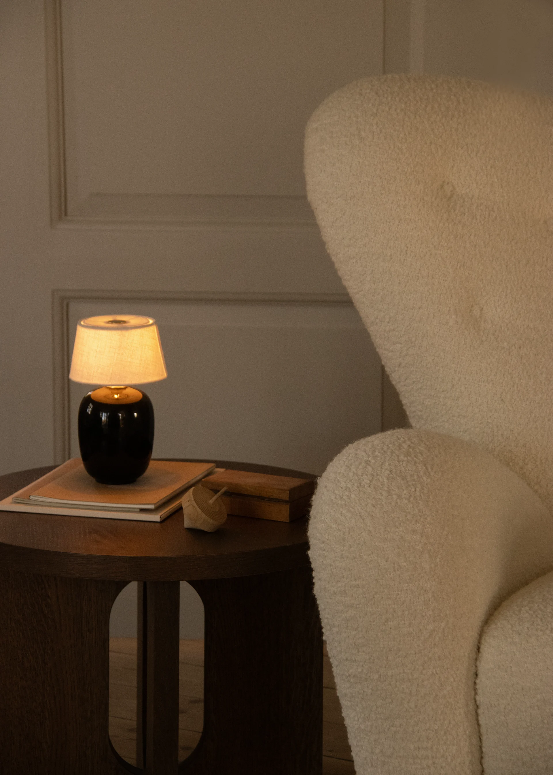 Coin chaleureux éclairé avec un fauteuil crème texturé et une table d'appoint en bois avec une lampe allumée, des livres et un jouet en bois.