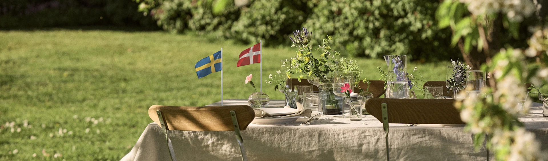 Table d'extérieur dressée avec des drapeaux suédois et danois, des fleurs sauvages et de la verrerie dans un jardin ensoleillé.