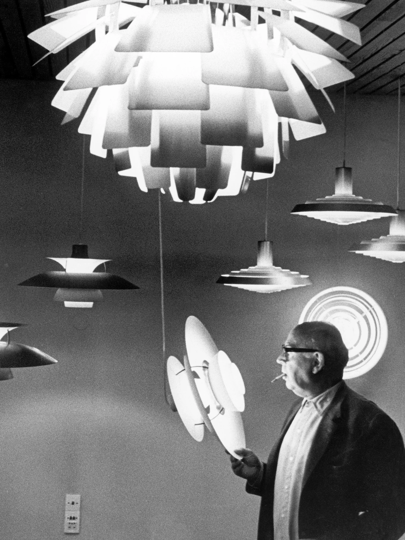 Homme à lunettes et cigarette inspectant plusieurs lampes modernes à couches en noir et blanc.