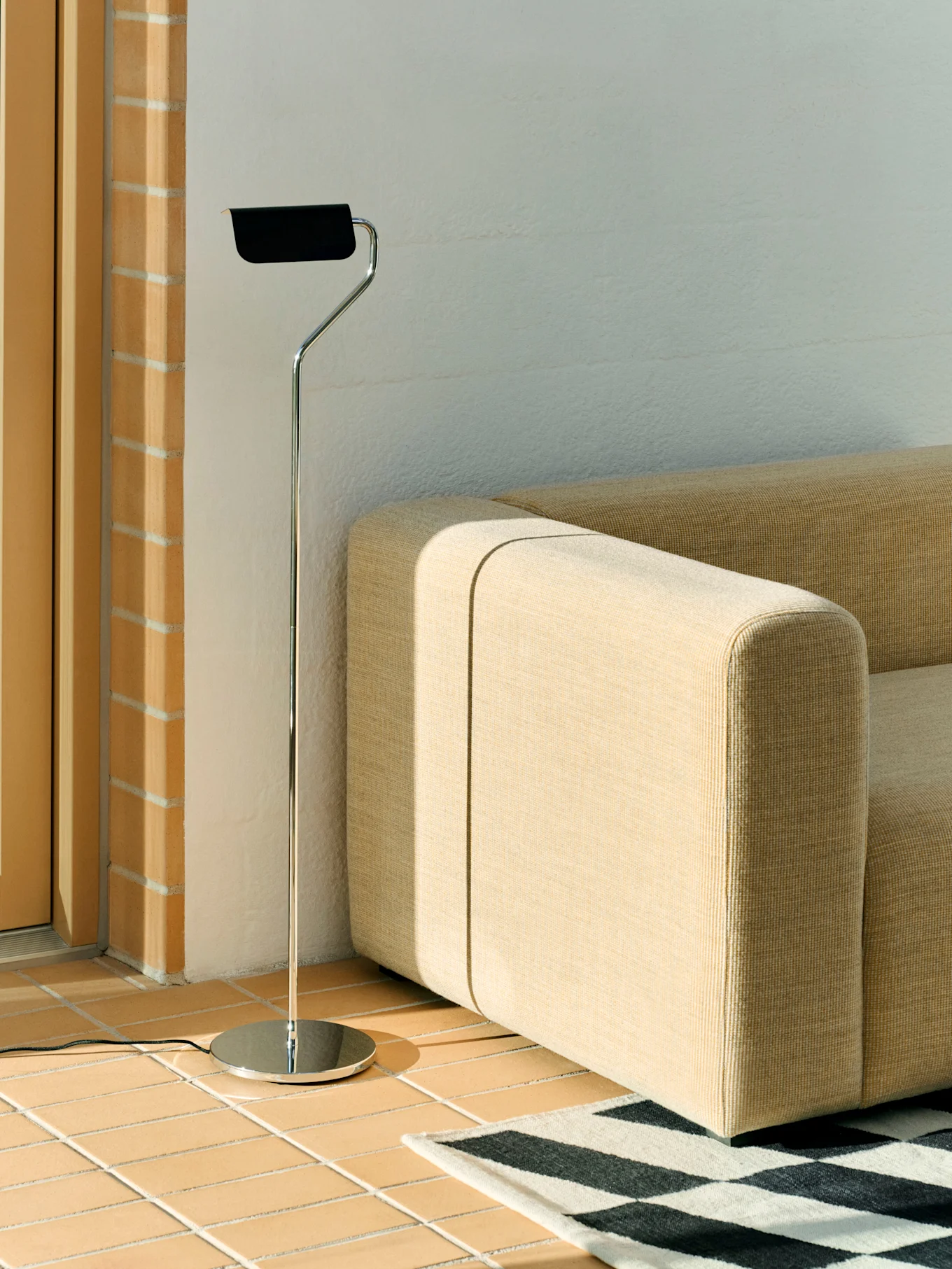 Lampadaire chromé avec abat-jour noir près d'un canapé modulaire beige et d'un tapis géométrique noir et blanc sur un sol carrelé.