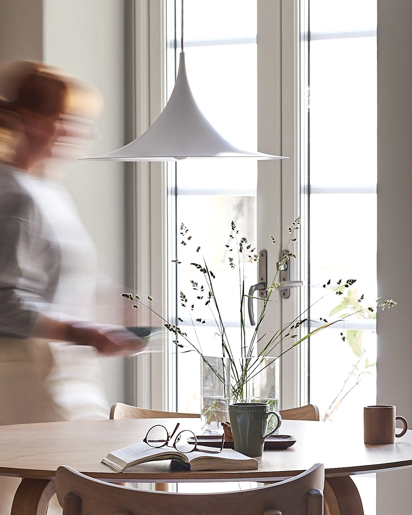 Une suspension blanche est suspendue au-dessus d'une table en bois clair avec un livre, des lunettes et des tasses. Une personne floue passe devant une fenêtre ensoleillée.