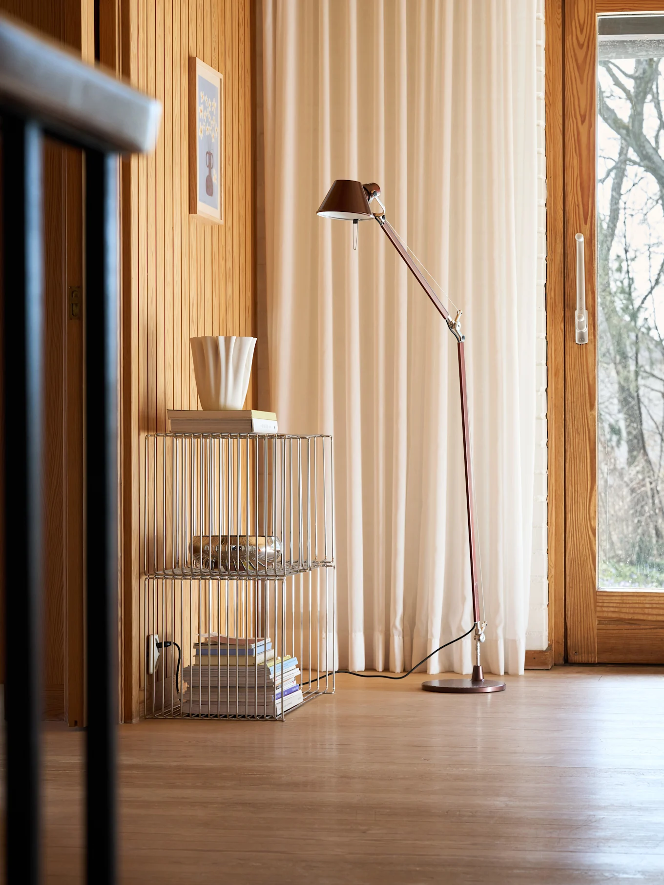 Une pièce chaleureuse avec des murs et un sol en bois, un lampadaire rouge, une étagère chromée avec des livres et un vase blanc cannelé, et une fenêtre avec des rideaux clairs.