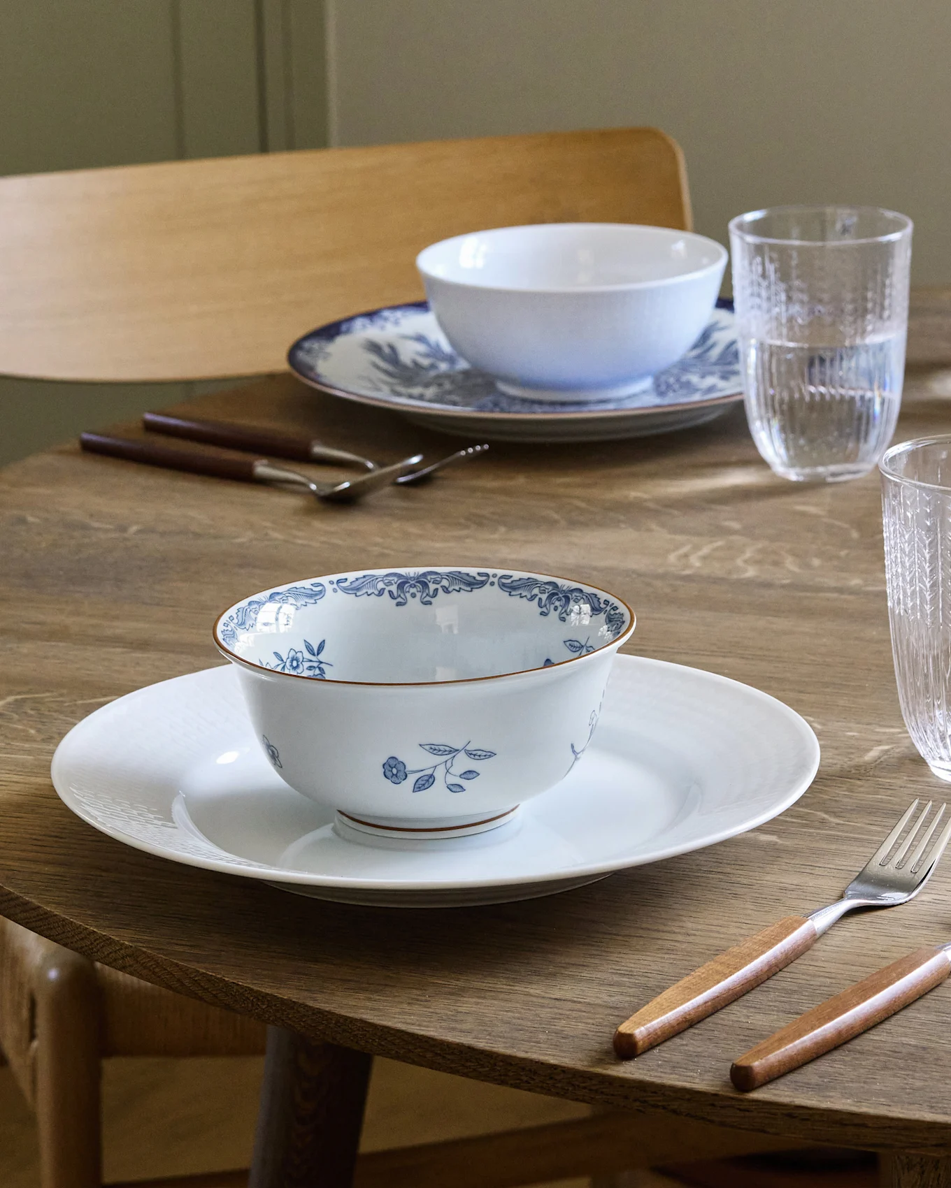 Table à manger dressée pour deux avec des bols à motifs blancs et bleus sur des assiettes blanches, des couverts à manche en bois et des verres d'eau.
