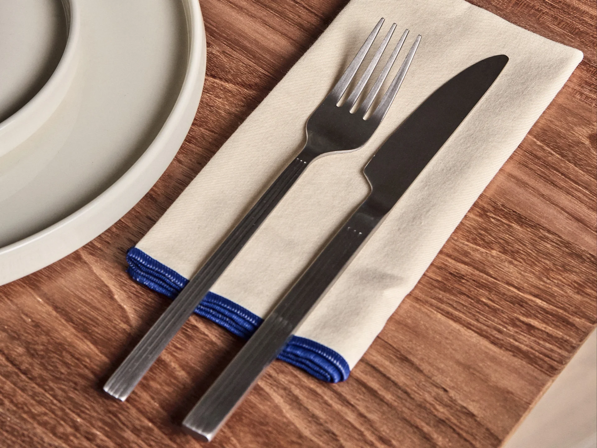 Fourchette et couteau sur une serviette crème avec une bordure bleue, à côté d'une assiette blanche sur une table en bois.