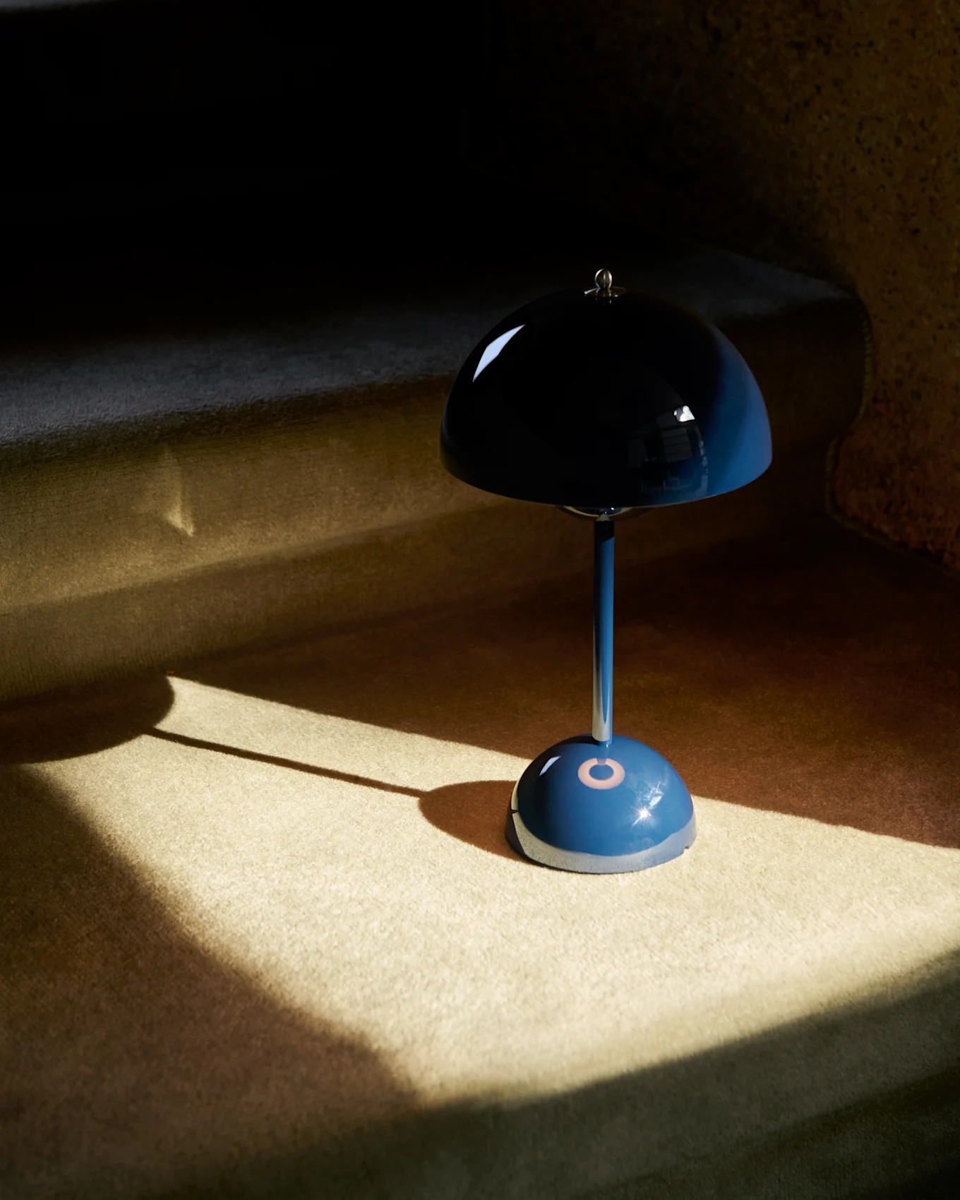 Une lampe de table bleue en forme de champignon avec un abat-jour réfléchissant posée sur un tapis, partiellement éclairée par le soleil.