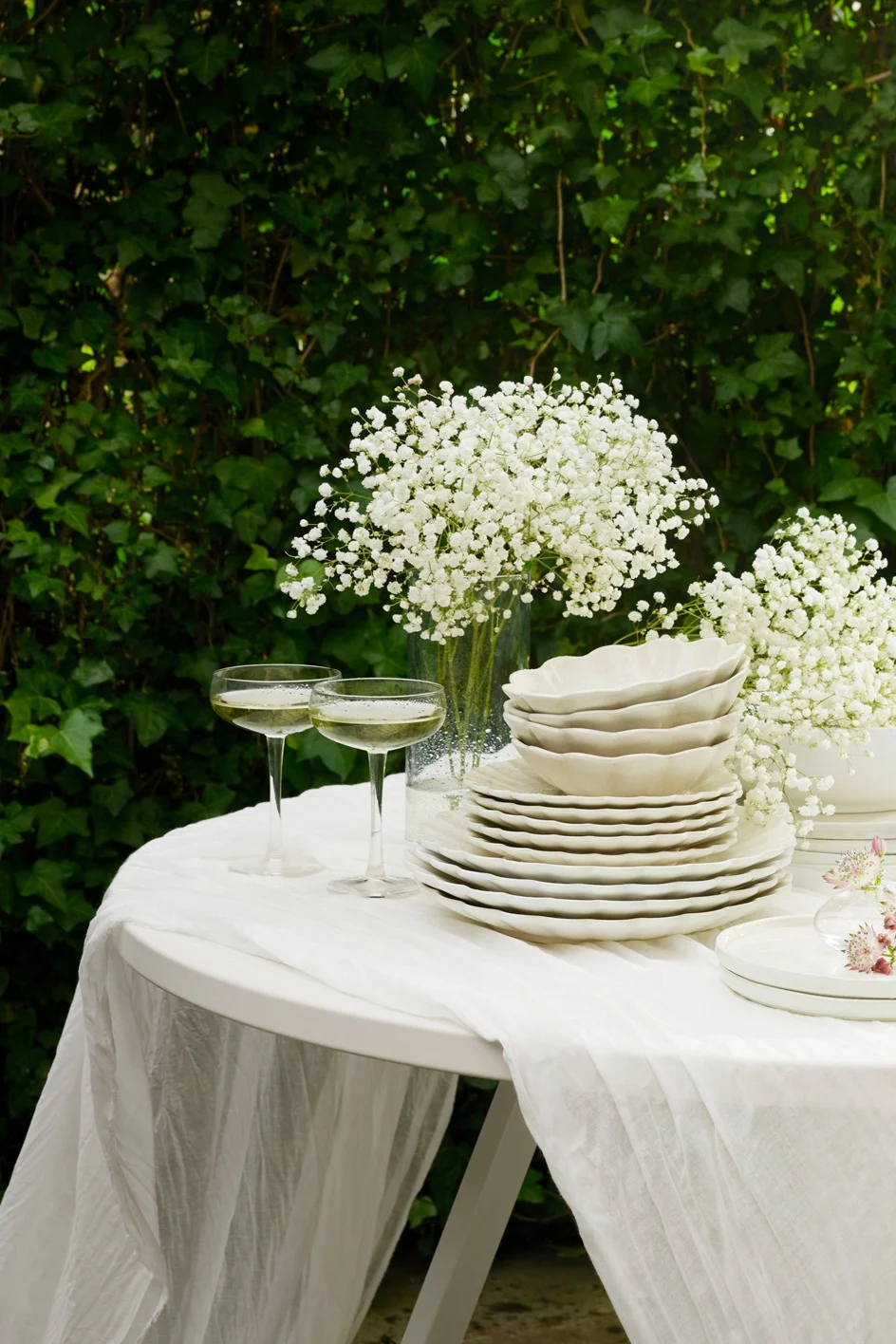 Table d'extérieur dressée avec vaisselle blanche, coupes de champagne et fleurs de gypsophile sur fond de feuillage vert.