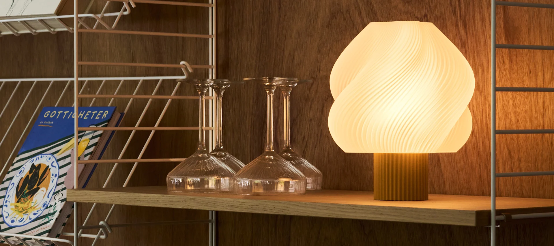 Une lampe ovale côtelée et illuminée et divers verres transparents sont posés sur un chariot de bar en bois devant un mur chaud et texturé.