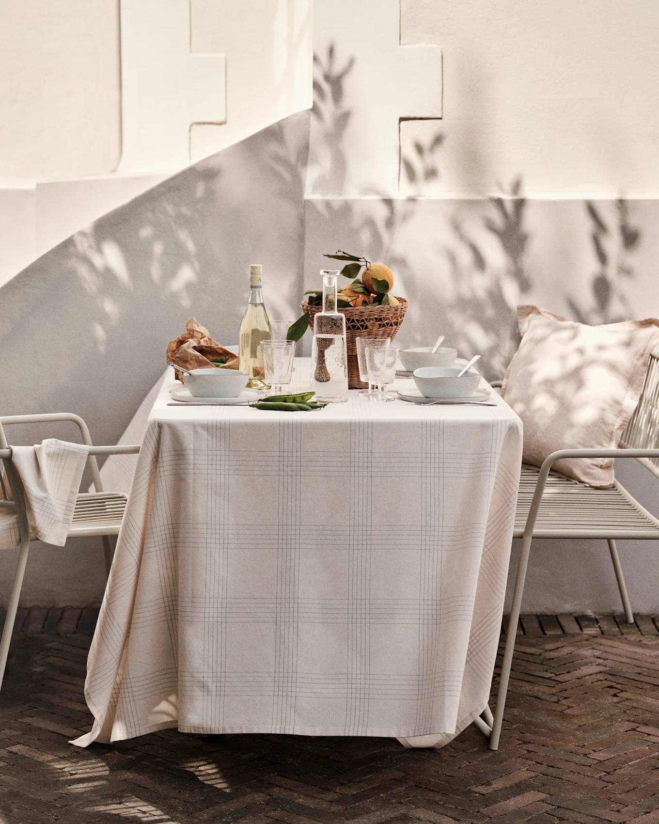 Table d'extérieur dressée avec nappe à carreaux, vin, eau, bols, panier d'oranges et petits pois sur un patio ensoleillé.