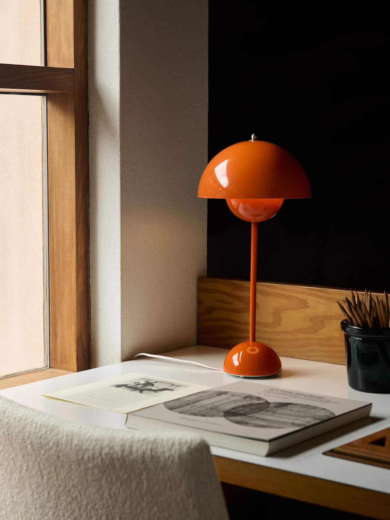Une lampe de table orange en forme de champignon sur un bureau blanc près d'une fenêtre, avec des livres et des crayons.