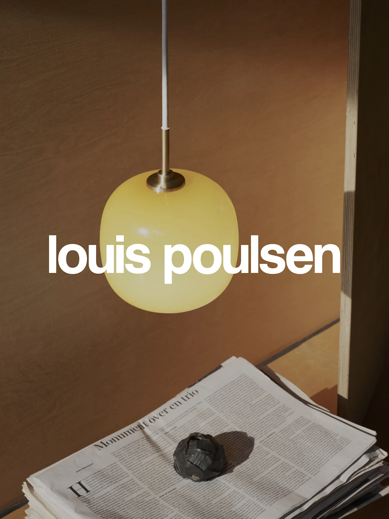 Logo Louis Poulsen superposé sur une scène chaleureuse avec une suspension sphérique jaune, des journaux empilés et un objet décoratif sombre.