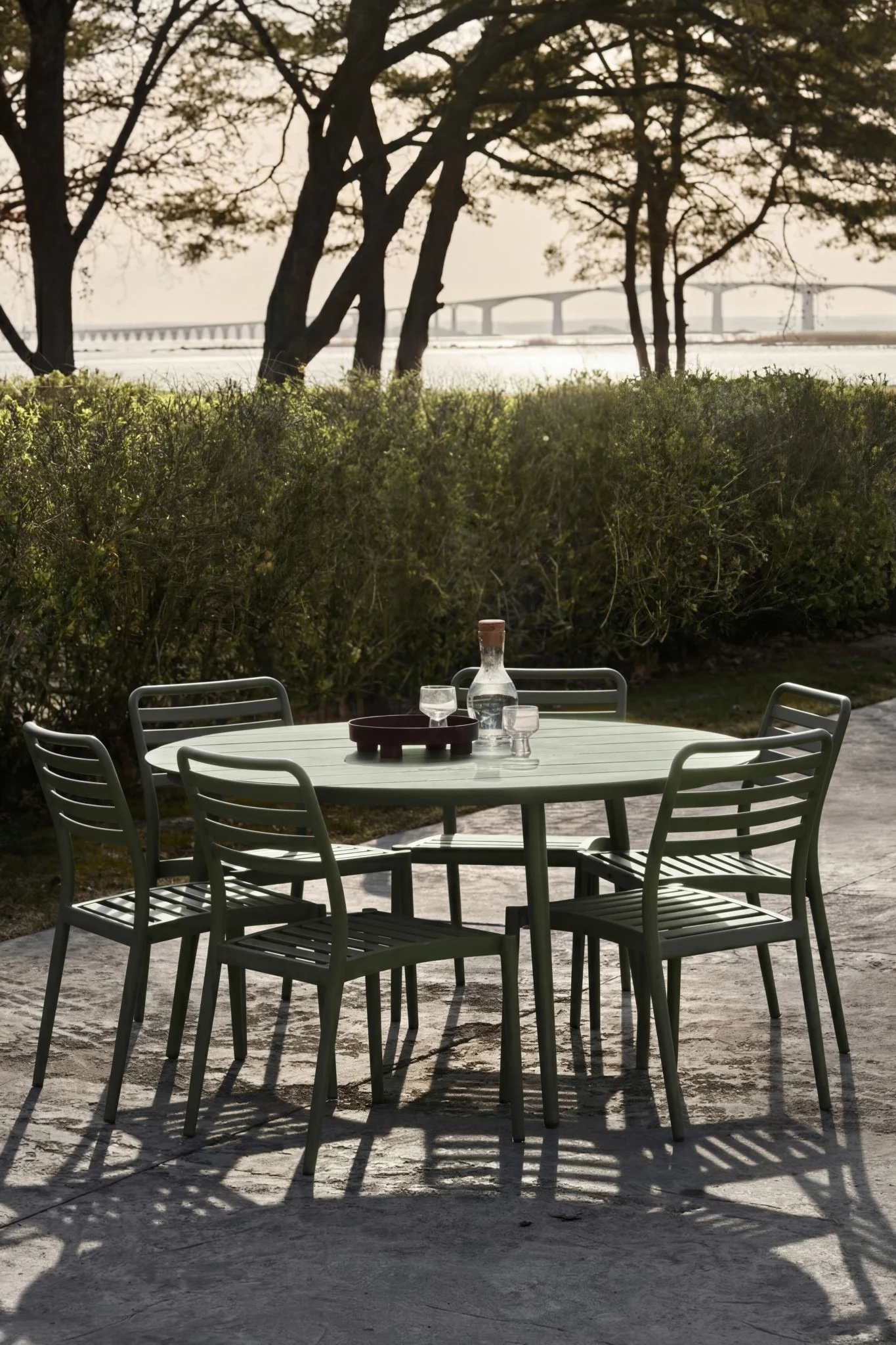 Ensemble de salon de jardin vert comprenant une table, cinq chaises, une bouteille d'eau et un verre sur la terrasse. Un pont enjambant un cours d'eau au loin.