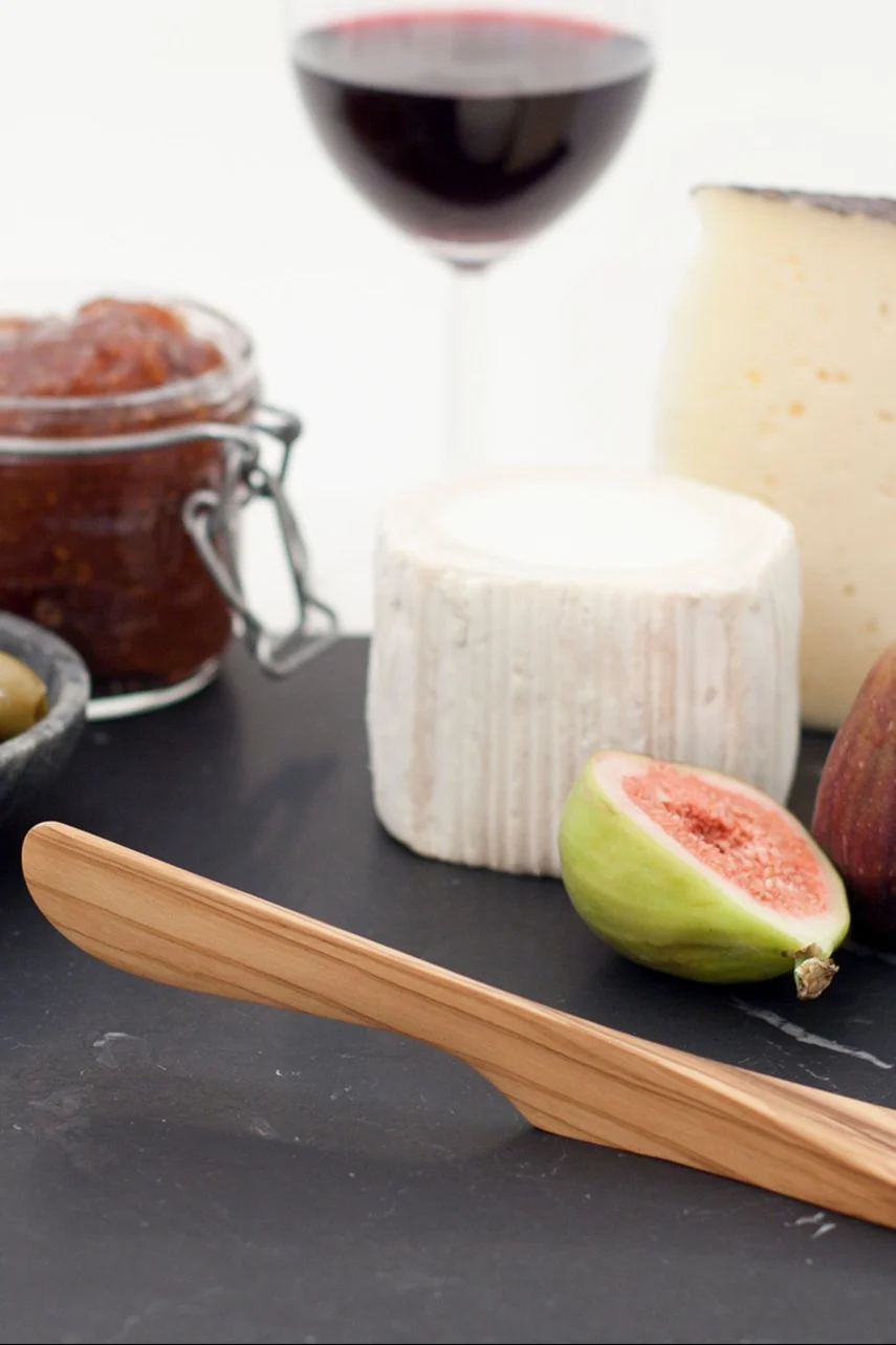 Un plateau de fromages avec un couteau en bois, deux fromages, une figue coupée en deux, de la confiture, des olives et un verre de vin rouge.