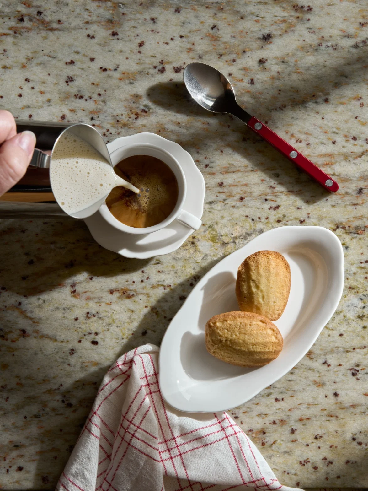 Main versant du lait mousseux dans une tasse de café à côté de madeleines sur un comptoir en granit.
