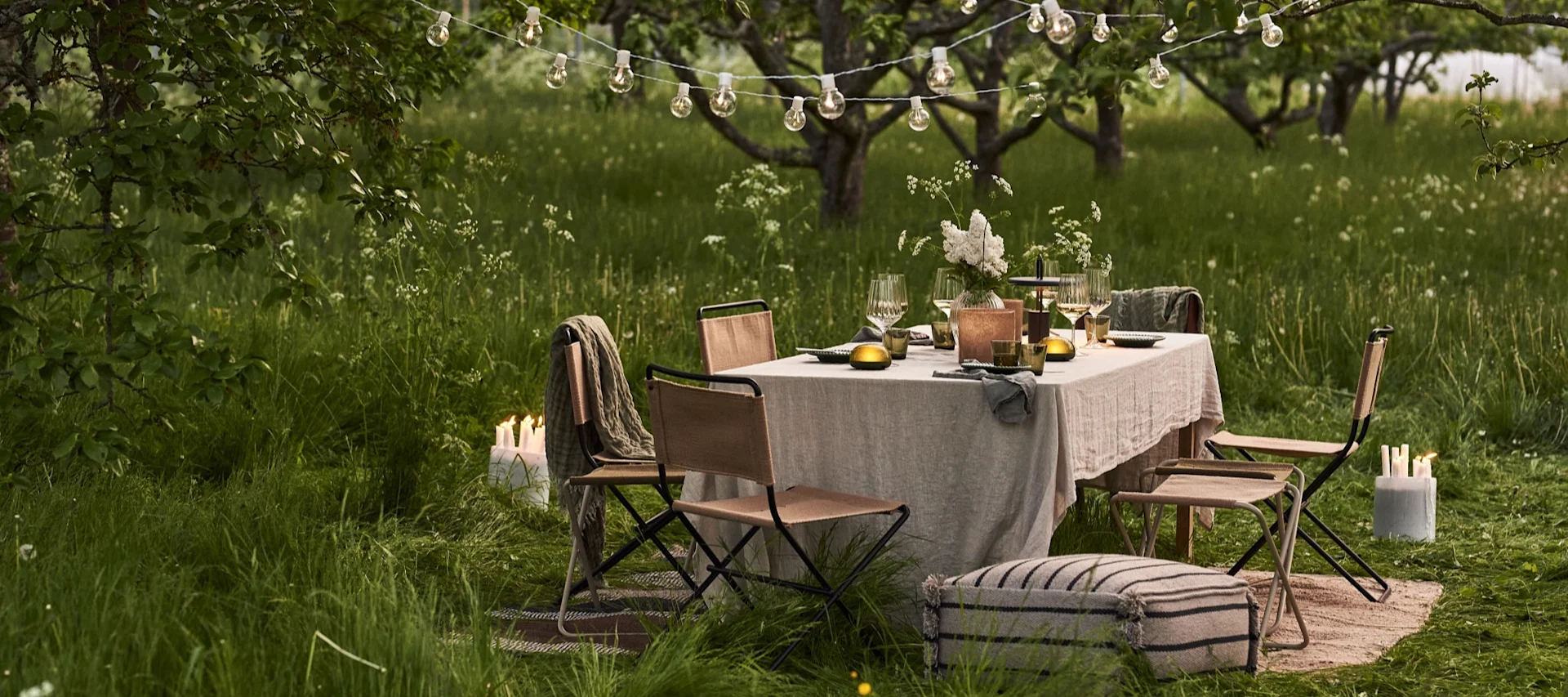 Ensemble de table à manger d'extérieur installé sous des guirlandes lumineuses dans un verger verdoyant au crépuscule, avec des chaises et des bougies d'ambiance.