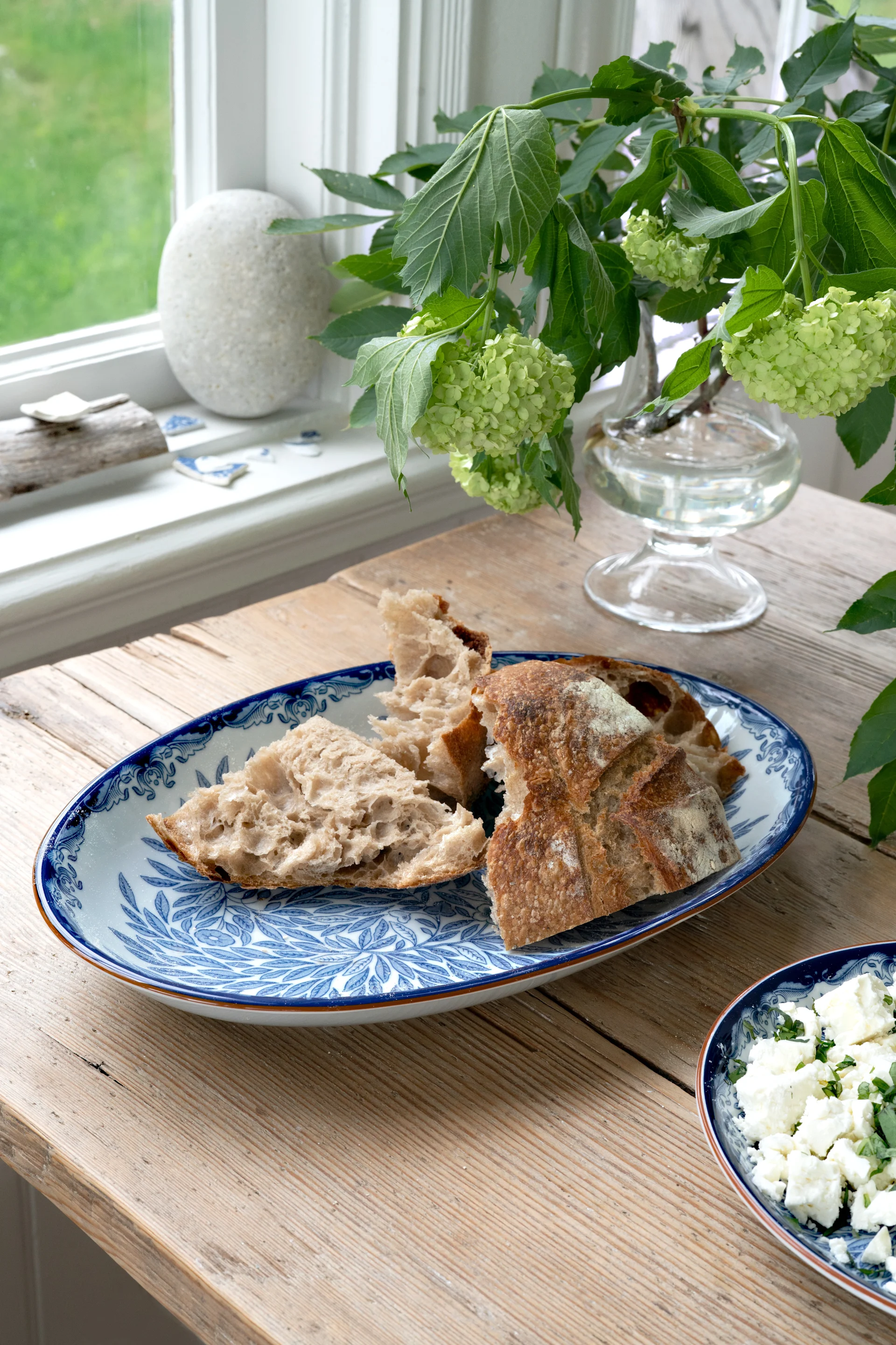 Morceaux de pain rustique déchirés sur une assiette à motifs bleus et blancs, avec du fromage émietté et des hortensias verts sur une table en bois.
