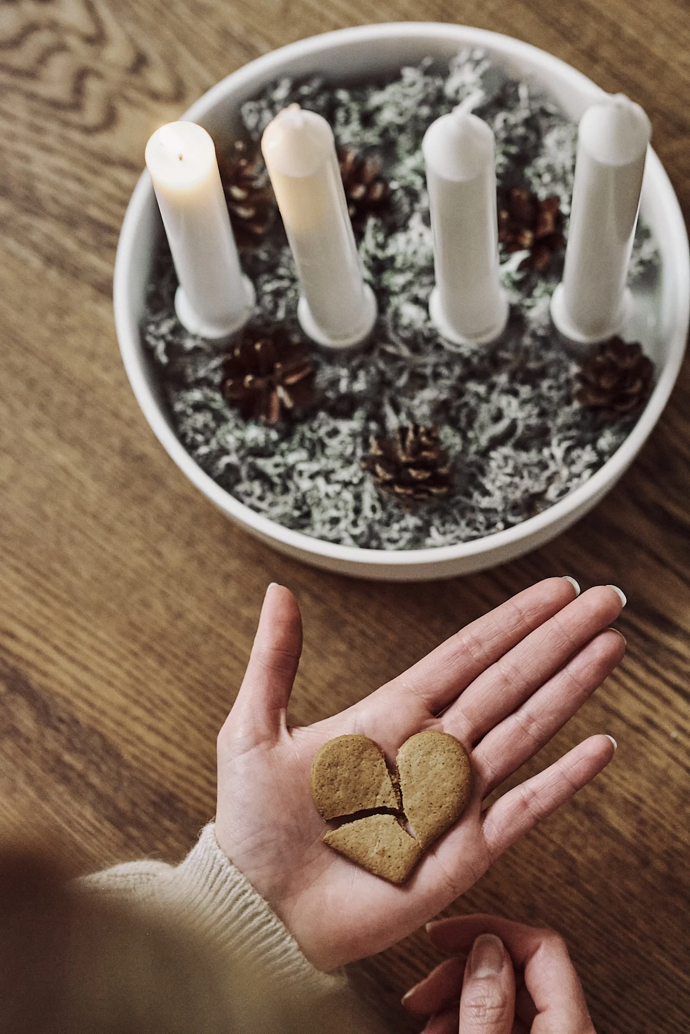 Sur une table se trouve le bougeoir de l'Avent Ernst décoré de pommes de pin et de branches de sapin avec 4 bougies, à côté duquel quelqu'un tient un biscuit cassé.