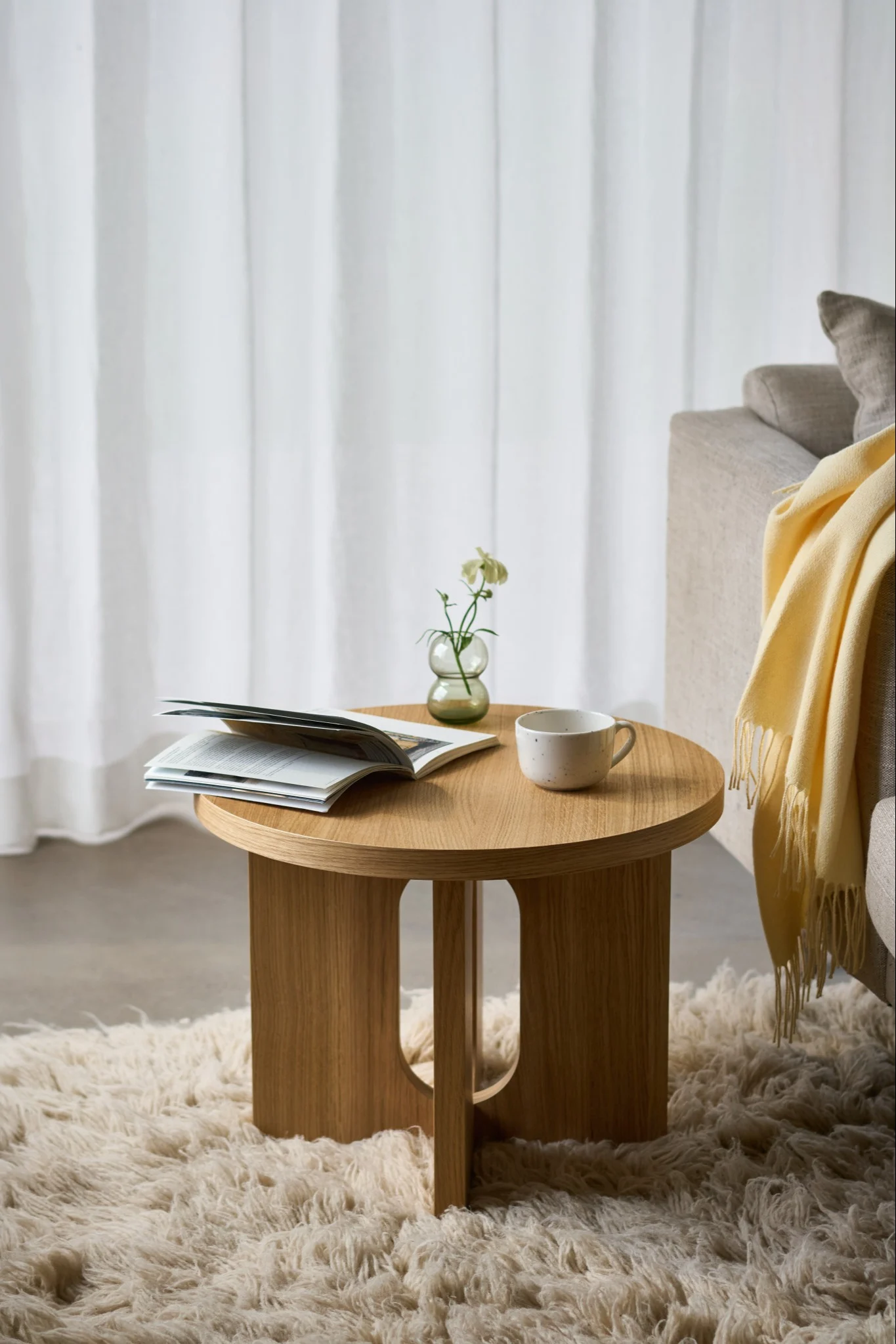 Une table d'appoint ronde en bois sur laquelle sont posés un livre ouvert, une tasse et un petit vase contenant une fleur blanche, le tout sur un tapis moelleux, à côté d'un canapé.
