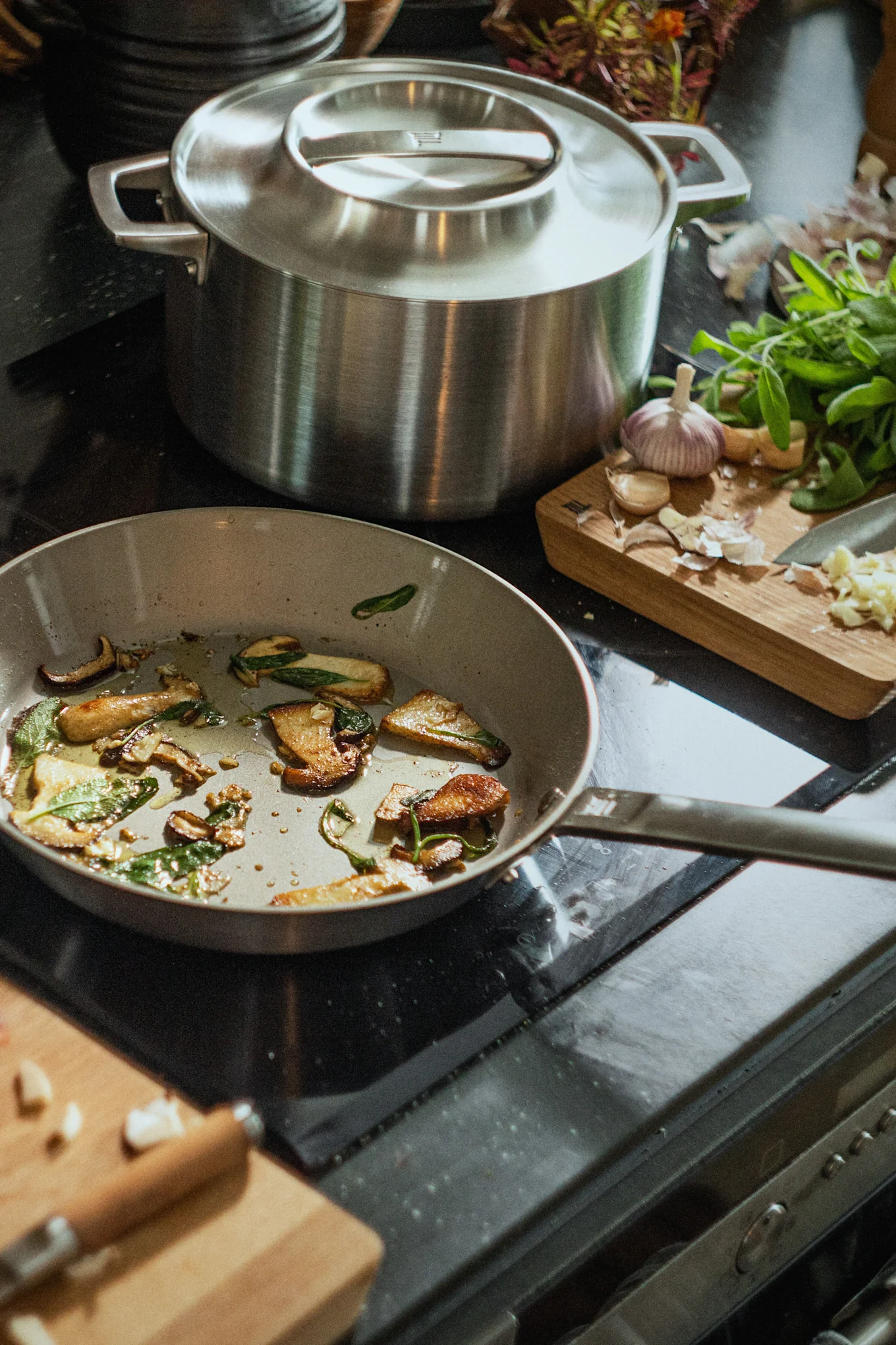 Une poêle avec des champignons et de la sauge sautés sur une cuisinière, à côté d'une grande casserole et d'une planche à découper avec de l'ail.